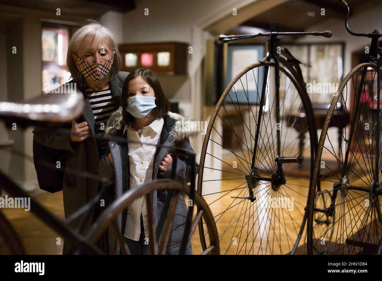 Tween schoolgirl and elderly female tutor in face masks viewing vintage bicycle in museum Stock Photo