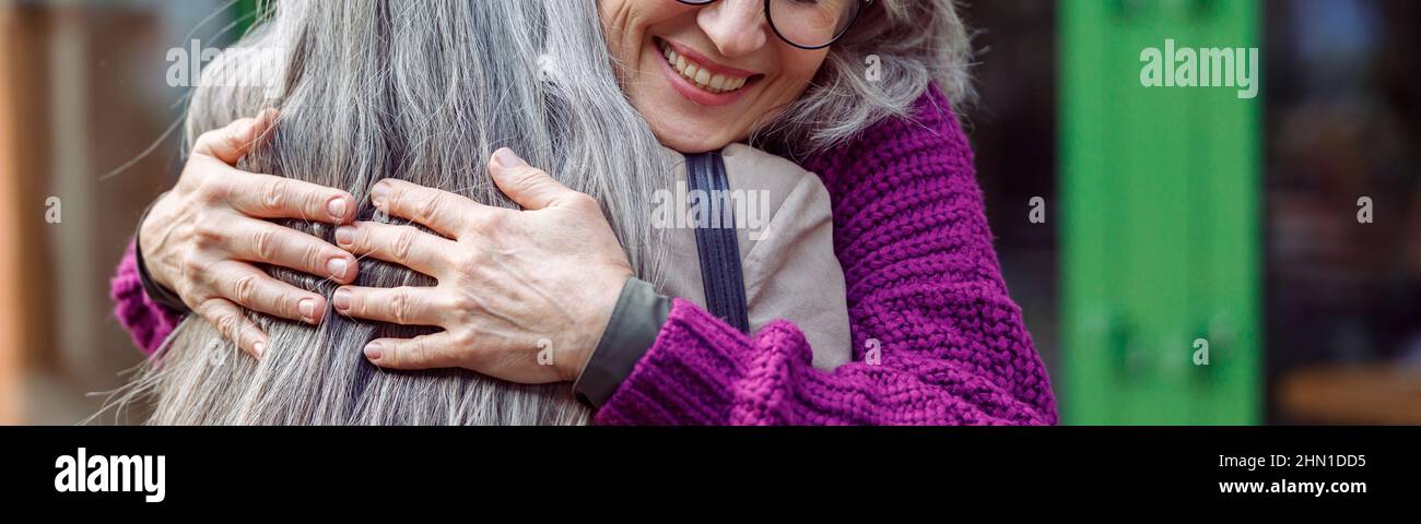 Smiling mature woman embraces female friend meeting on modern city street Stock Photo