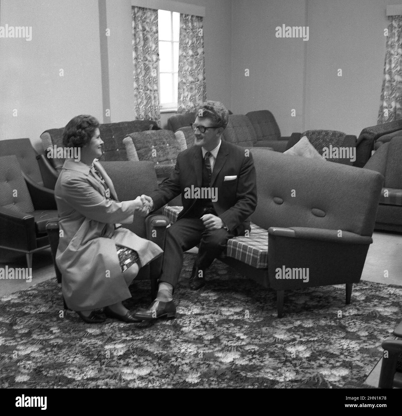 1960s, historical, inside a furniture shop, a lady shaking the hands of a suited salesman for his help after buying a new settee and lounge chairs, known as a 'three piece suite'. Stock Photo