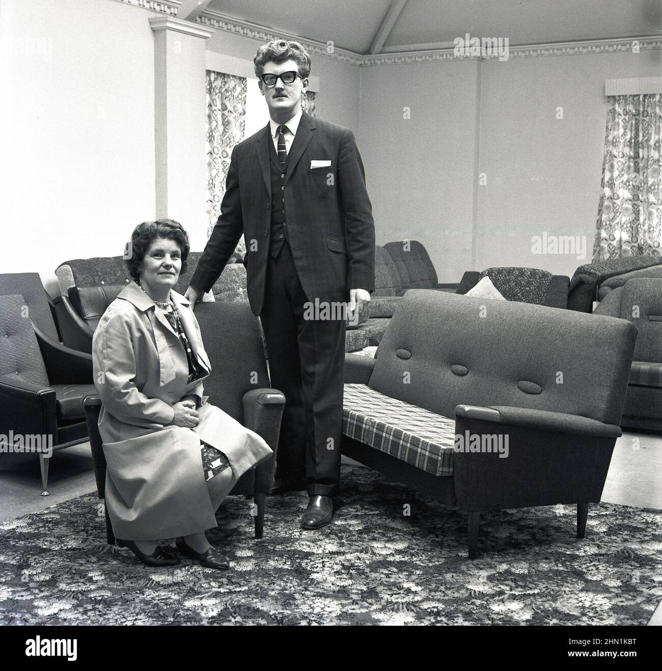 1960s, historical, inside a furniture shop, a lady with a suited salesman pose for their photo after buying a new settee and lounge chairs, ofen referred to as a 'three piece suite'. Stock Photo