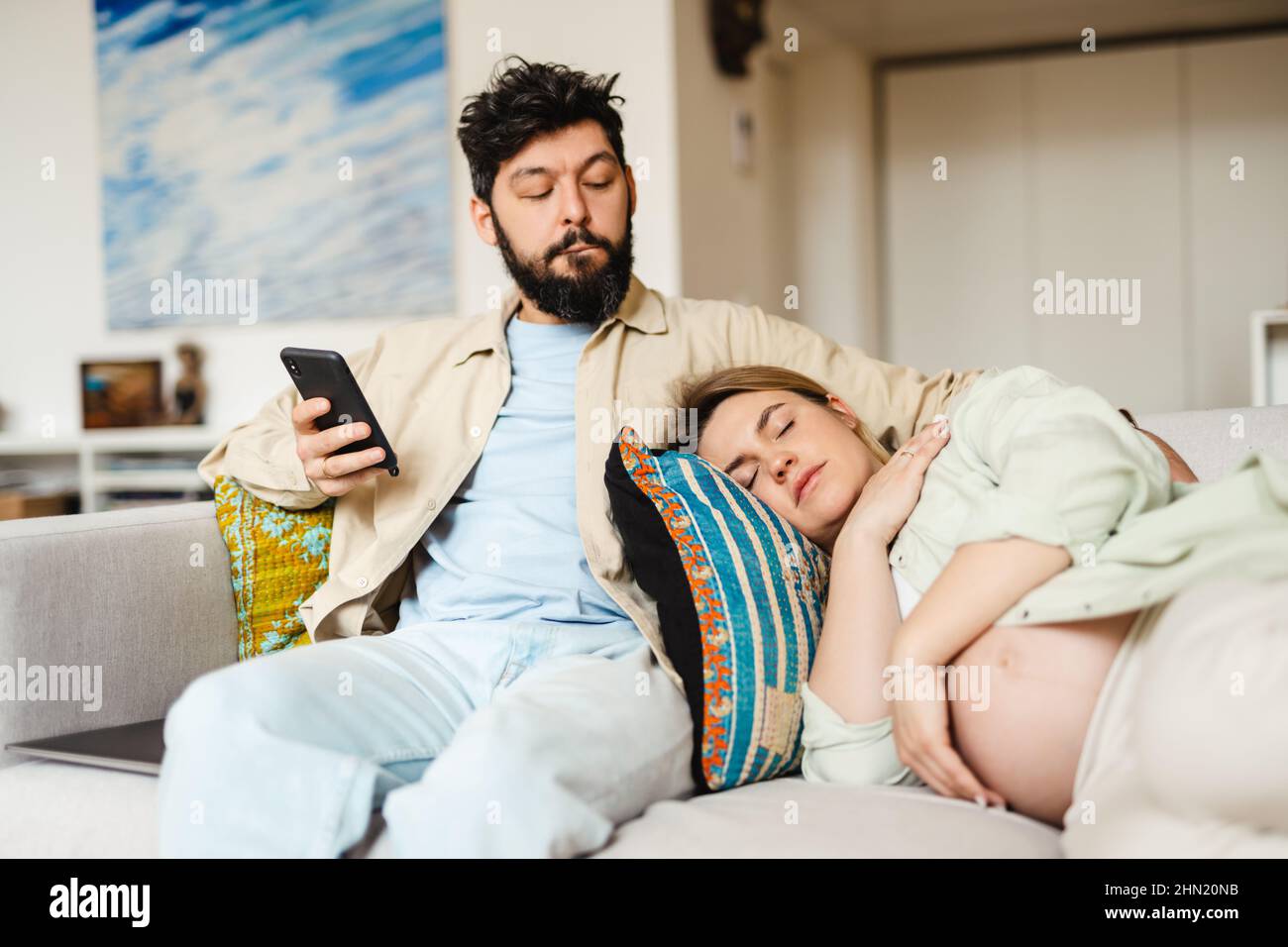 Bearded man using cellphone while his pregnant wife sleeping on couch