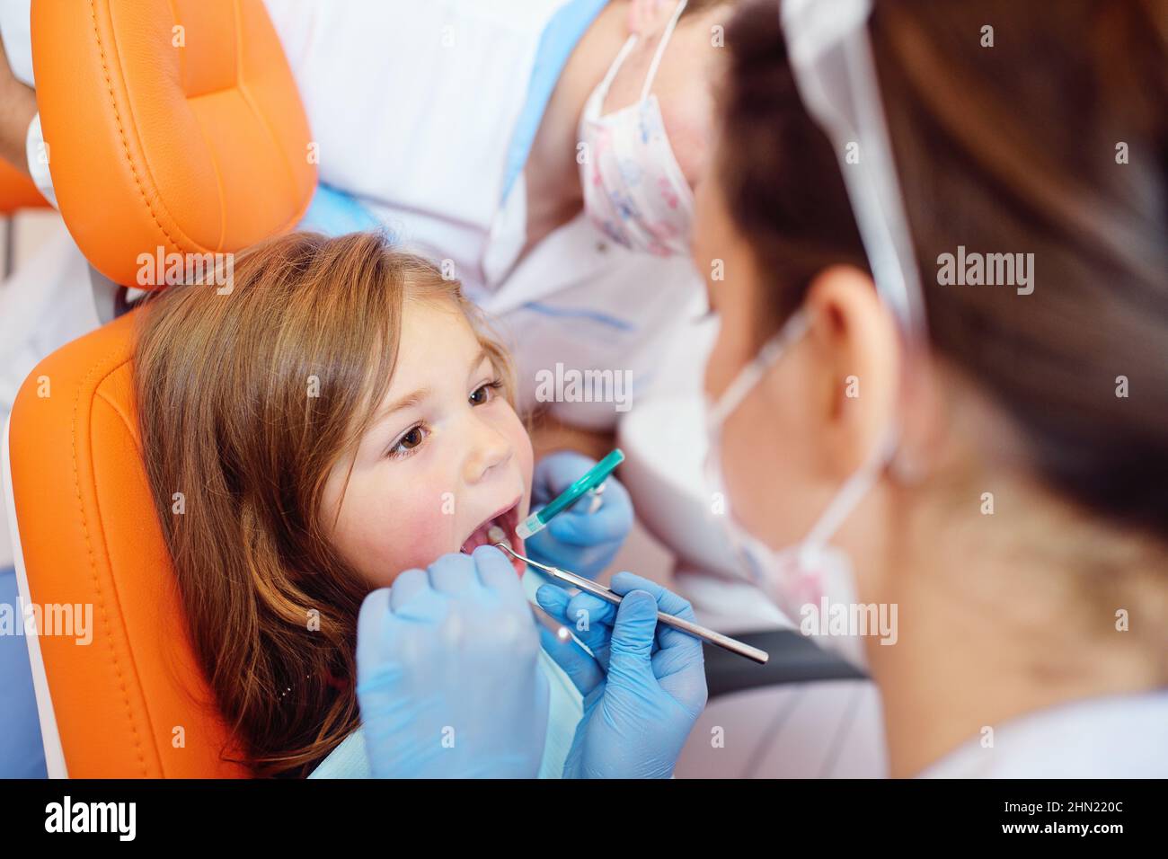 young dentists, a man and a woman, examine the teeth of a child's patient - a little pretty girl who is sitting in an orange dental chair. Stock Photo