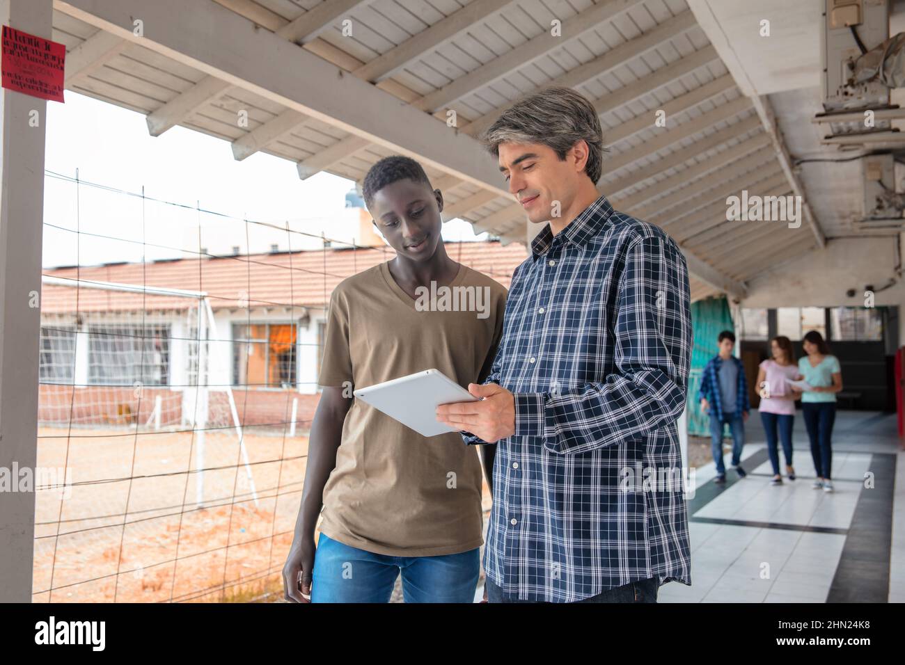 Hight school african student talking with his teacher in the hallway, other students talking in the background Stock Photo