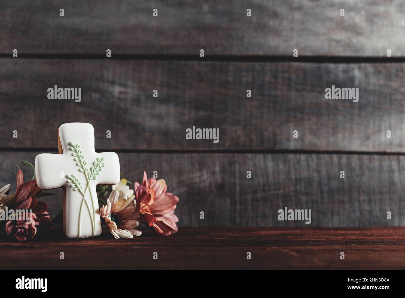 Easter holiday conceptual background on rustic wooden boards. Photo of white gingerbread cookie Cross shape, flowers bouquet on table top. Card with copy space to place text. Minimal concept Stock Photo