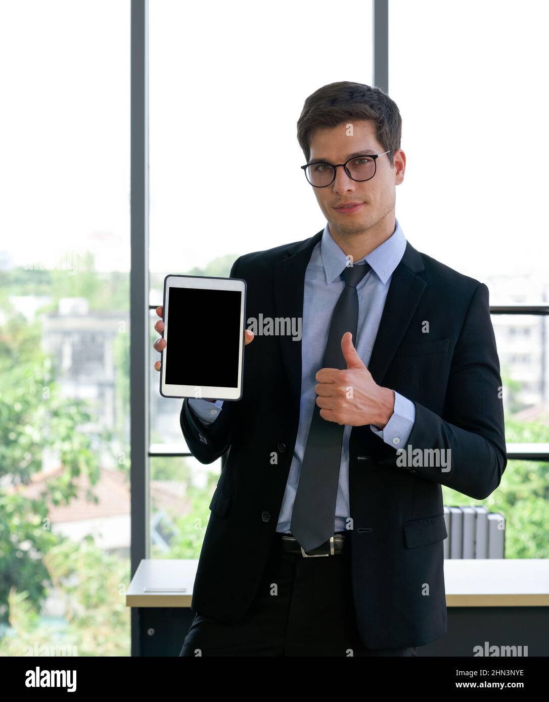 Business people in suits and ties raise finger thumbs up while holding blank screen tablet computer. Morning work atmosphere in a modern office. Stock Photo