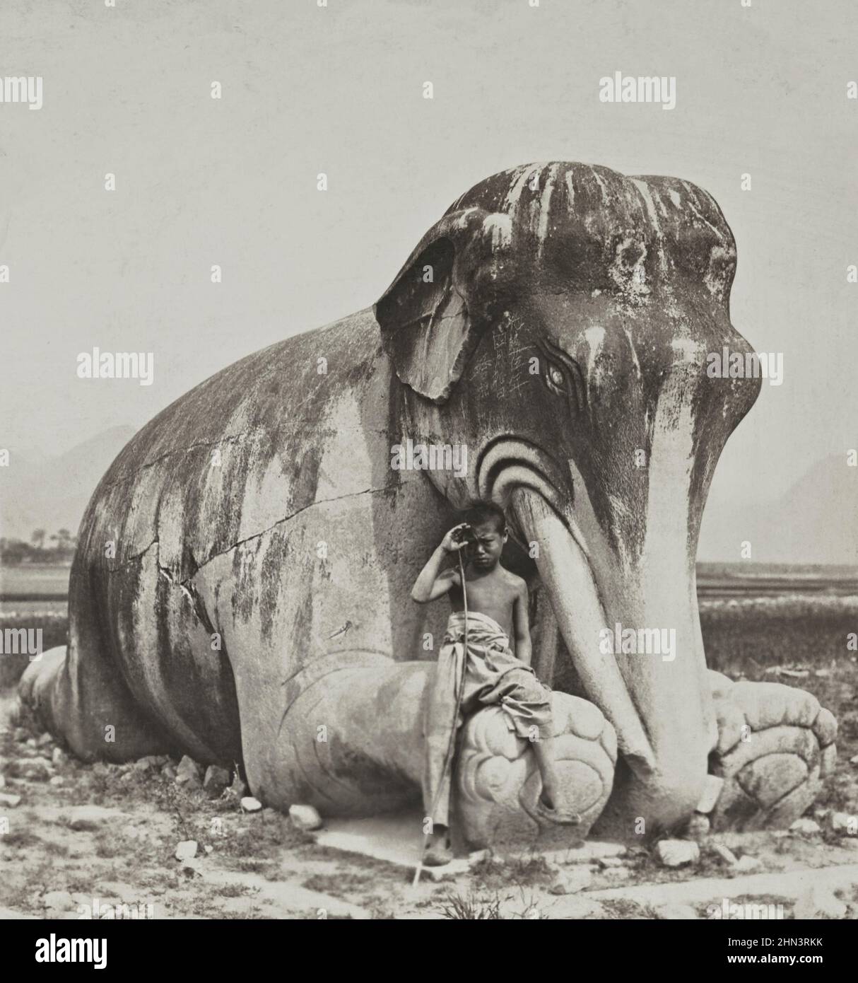 Vintage photo of young Chinese herd near ancient Chinese sculpture of a kneeling stone elephant. The Ming Tombs. North China. 1902 Stock Photo