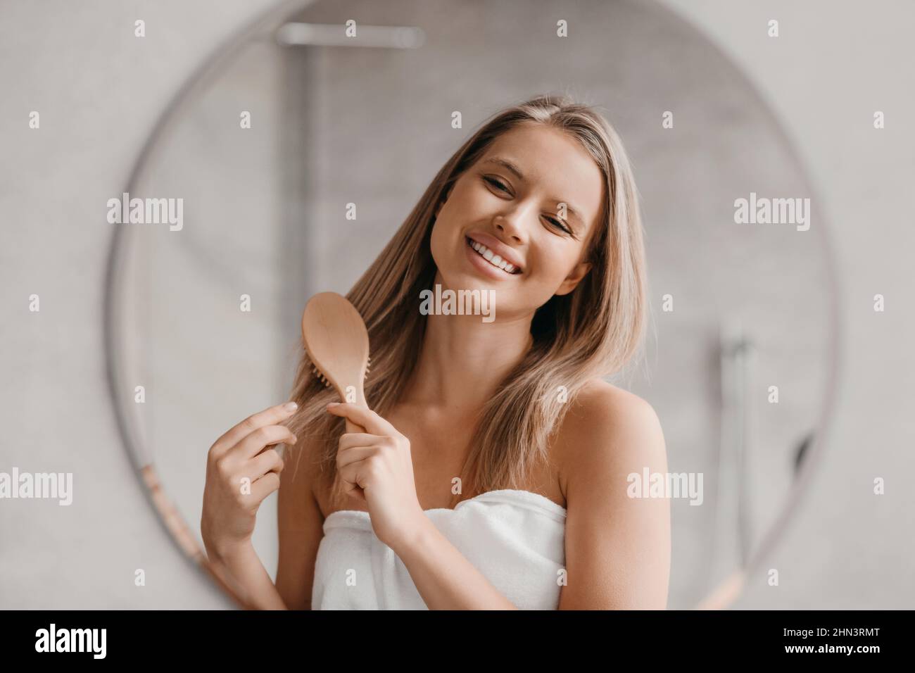 Haircare concept. Pretty lady brushing her beautiful hair with comb while looking at her reflection in mirror Stock Photo