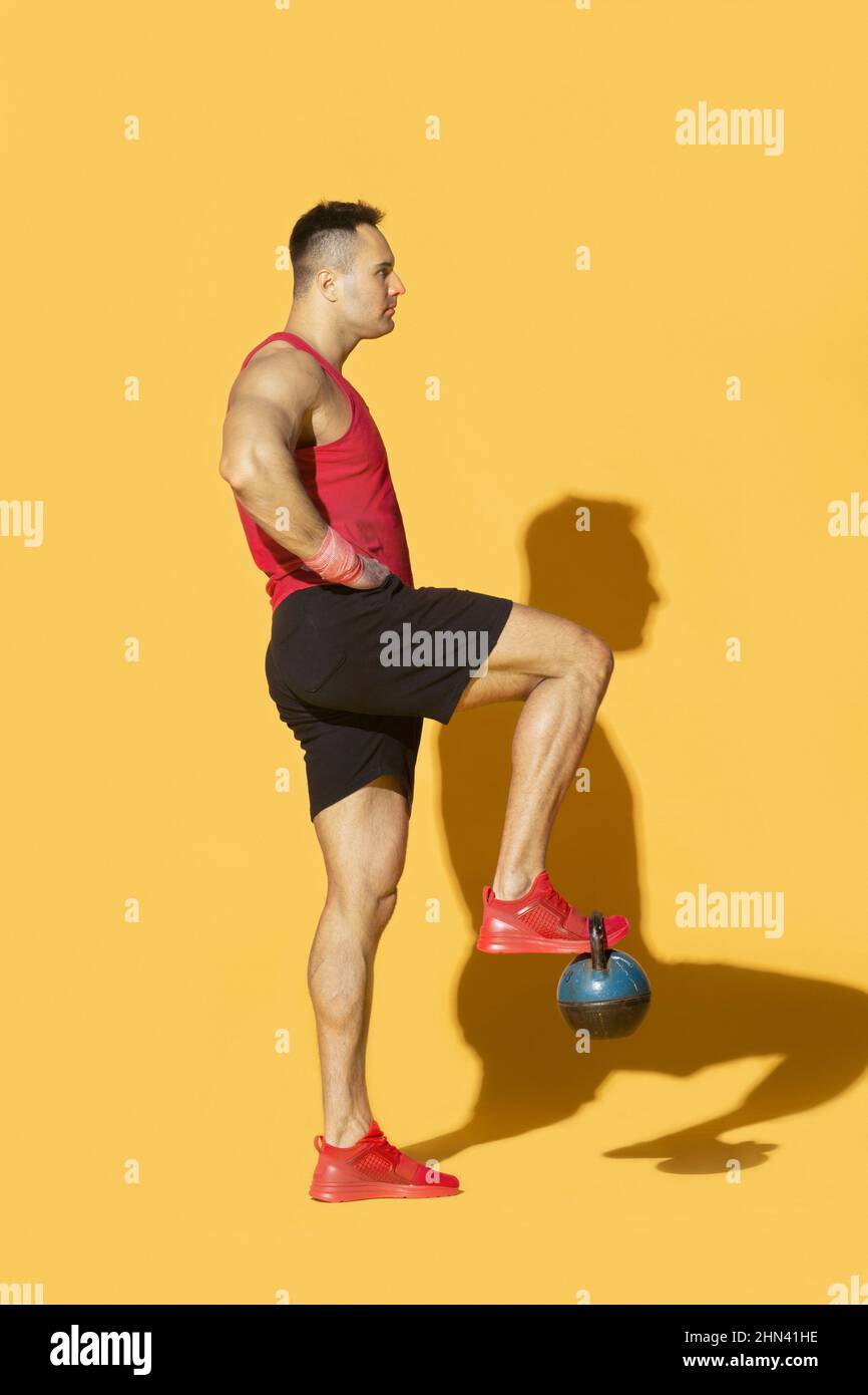 Athletic man in sportswear lifting kettle bell with foot against yellow background Stock Photo