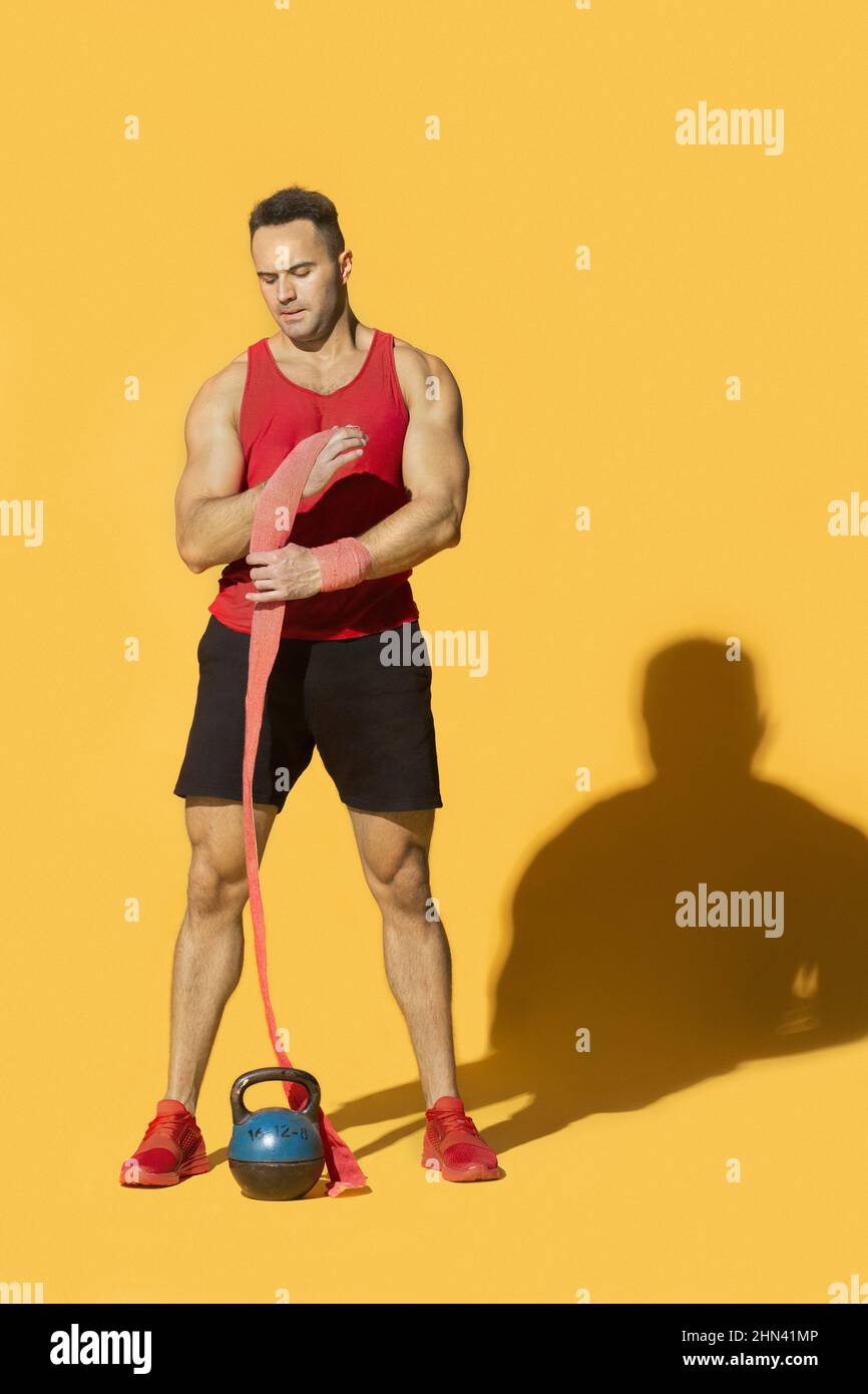 Athletic, strong man with kettle bell wrapping wrist with bandage against yellow background Stock Photo