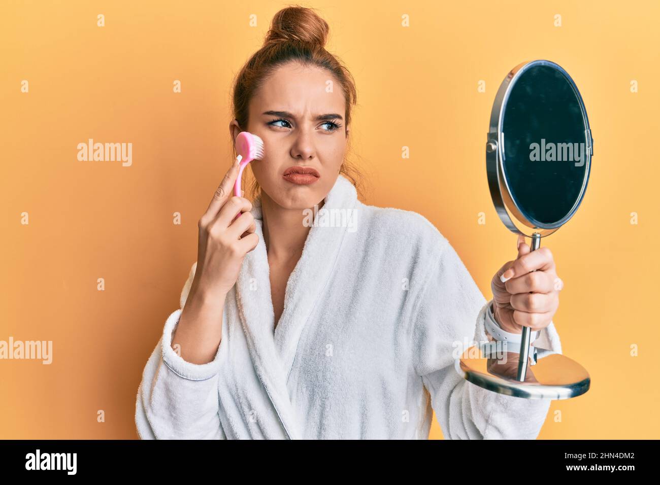 Young blonde woman wearing robe holding makeup remover brush skeptic and nervous, frowning upset because of problem. negative person. Stock Photo