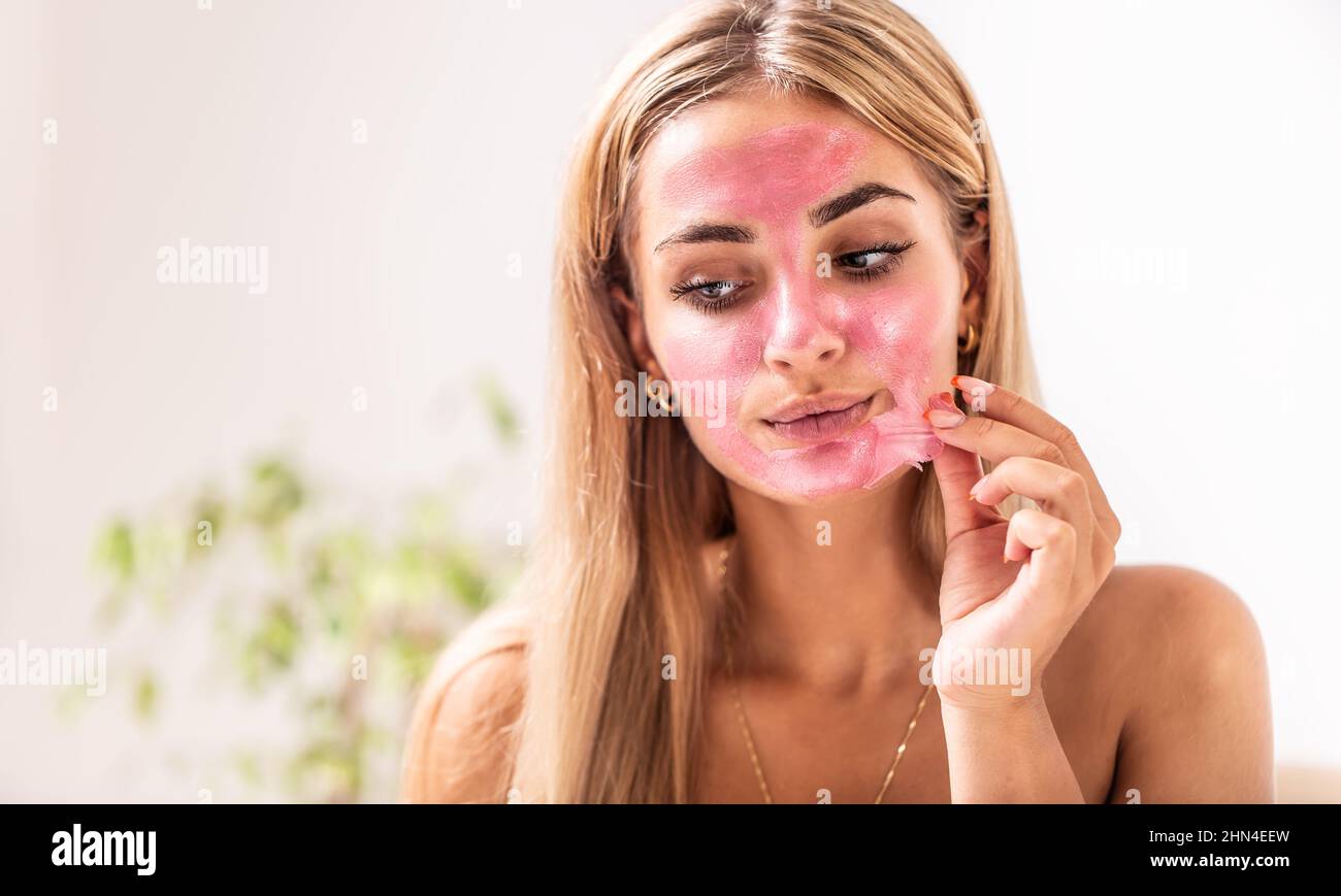 Caucasian blonde girl applies pink peeling mask on her face indoors. Stock Photo