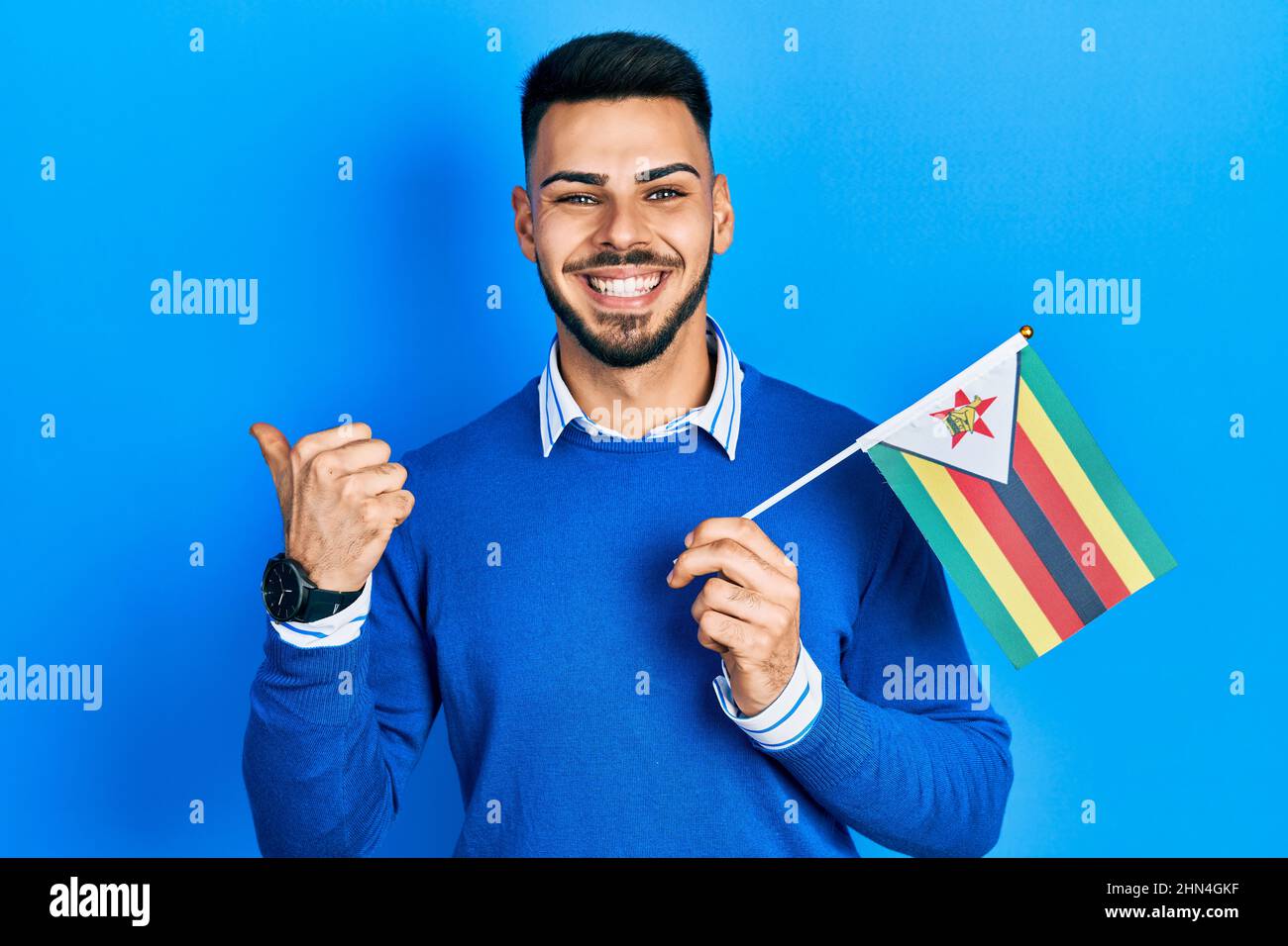 Young hispanic man with beard holding zimbabwe flag pointing thumb up to the side smiling happy with open mouth Stock Photo