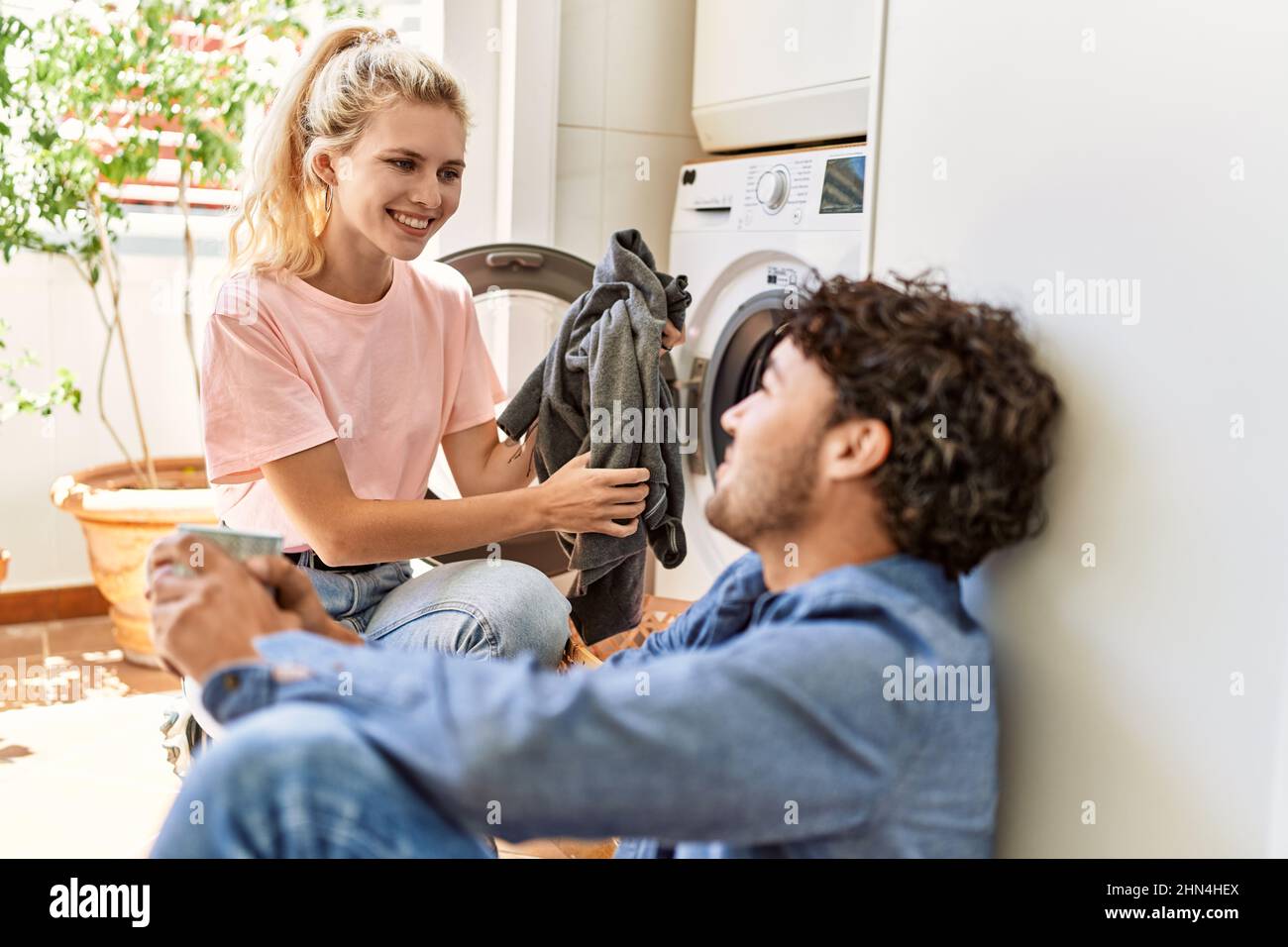 Young couple smiling happy drinking coffee while doing laundry at home ...