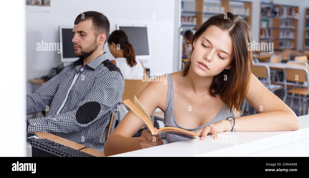 Girl reading book and using pc in college library Stock Photo - Alamy