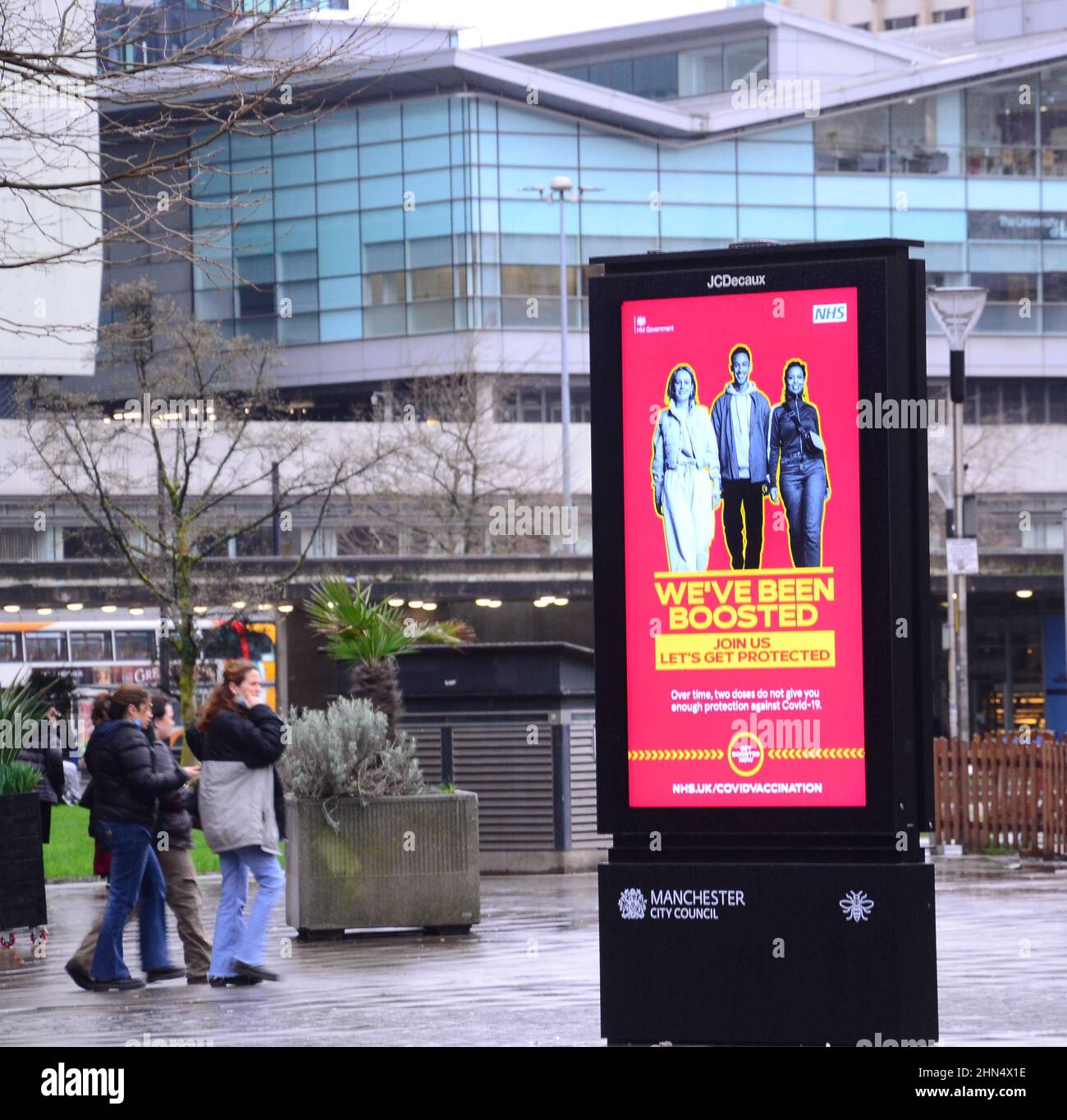 People walk past an NHS and HM Government 'We've Been Boosted' electronic sign to advertise the booster vaccine to combat Covid-19 in Manchester, Greater Manchester, England, United Kingdom. The number of Covid-19 or Coronavirus or Corona infections is continuing to fall in Greater Manchester. 12,364 people tested positive in the area in the week ending February 8th, 2022, a 35 per cent fall on the prior  week. Stock Photo