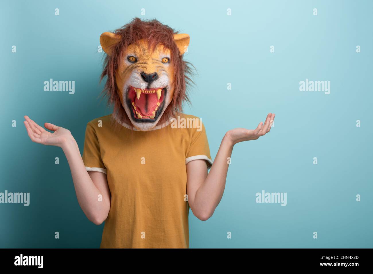 Confused young woman in lion mask standing with raised open hands and shrugging shoulders Stock Photo