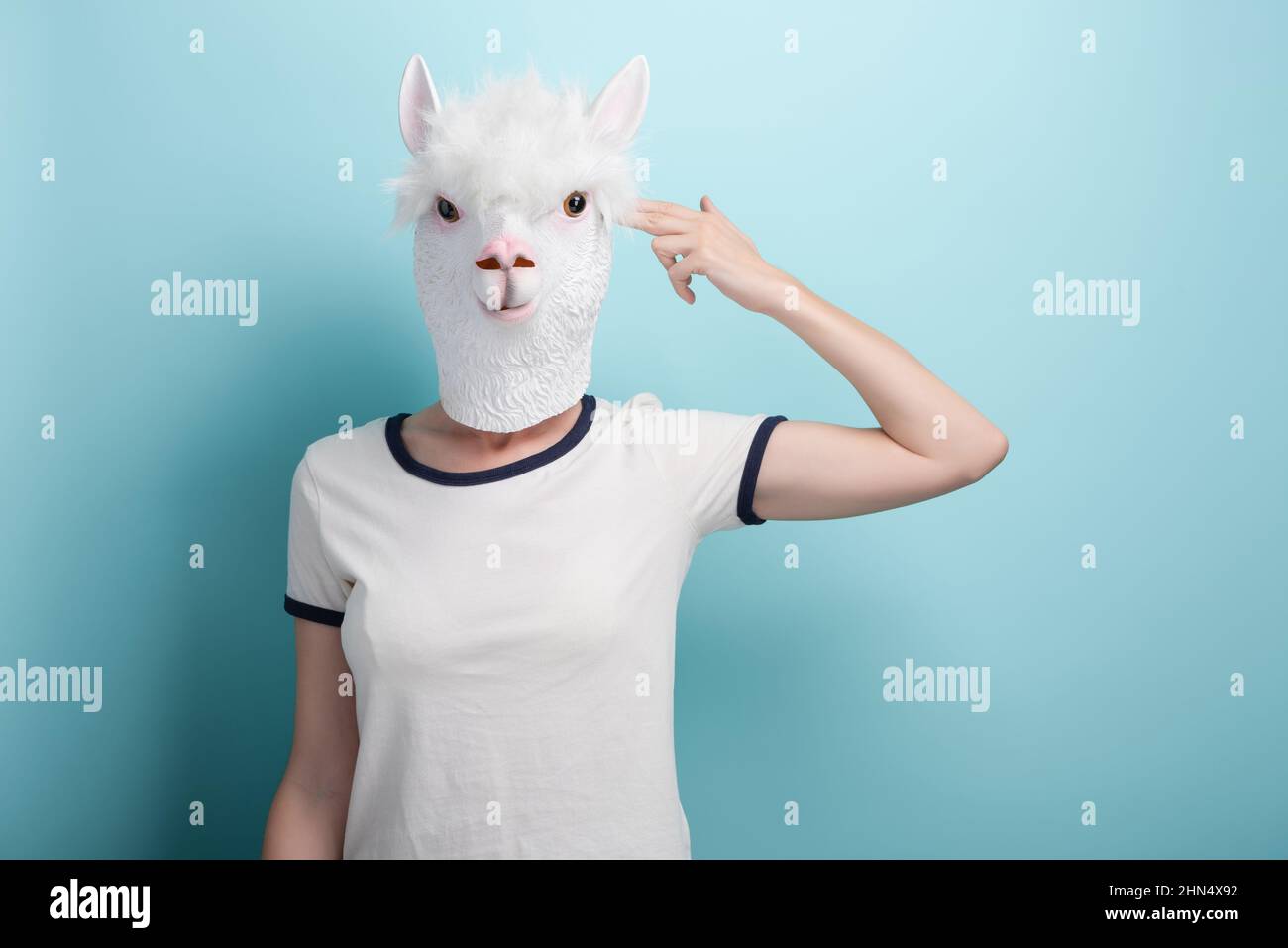 Young woman in alpaca mask pointing fingers to head in gun hand gesture imitating suicide, isolated on blue background. Stock Photo