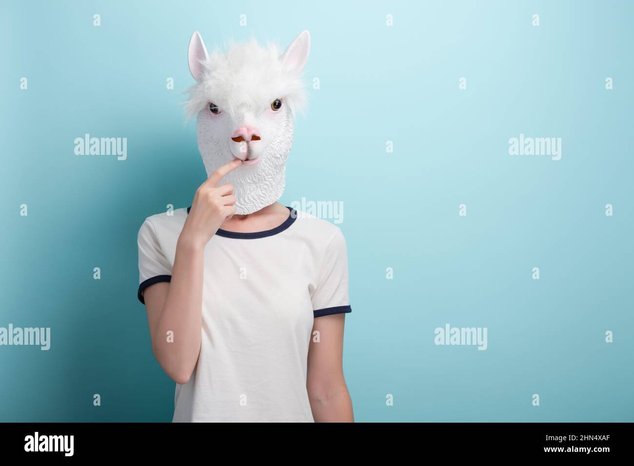Young woman in alpaca mask with index finger in mouth, thinking or in doubt, isolated on blue background with copy-space. Stock Photo
