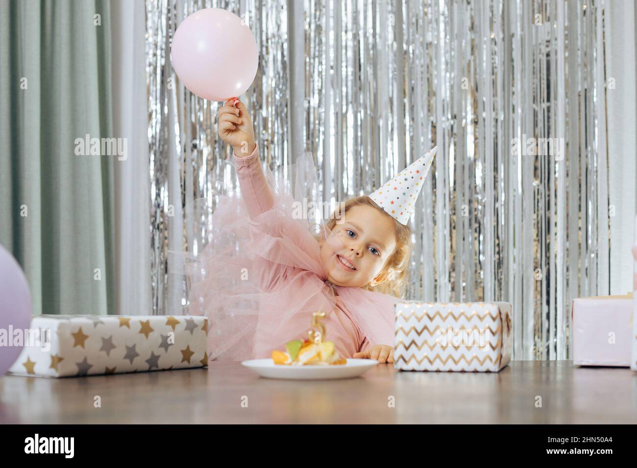 Cute little smiling girl with short curly hair and stars on face in pink poofy dress lift pink balloon near fruit cake. Stock Photo