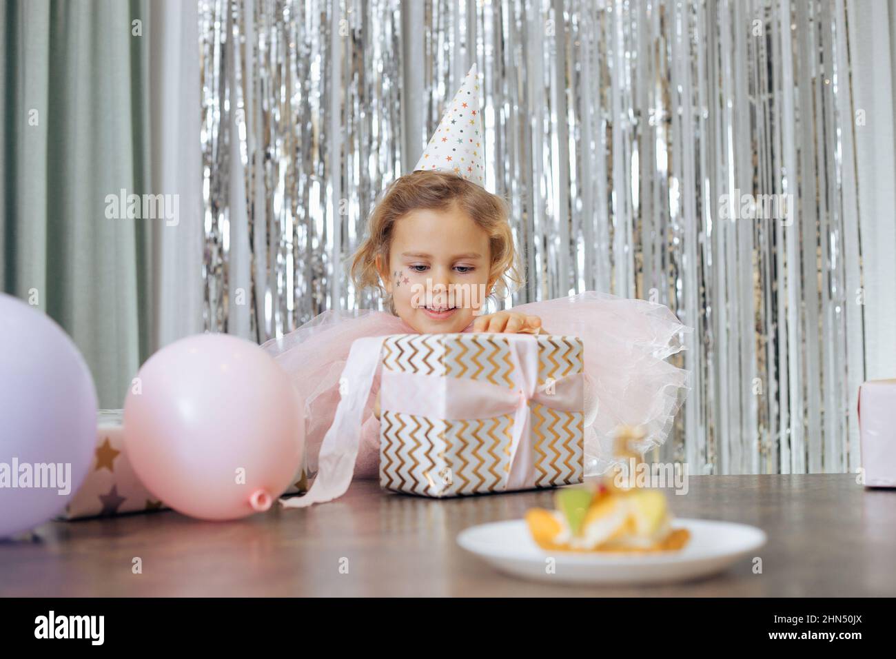 Cute little smiling girl with short curly hair and stars on face in pink poofy dress, cap unwrap gift box near balloons. Stock Photo