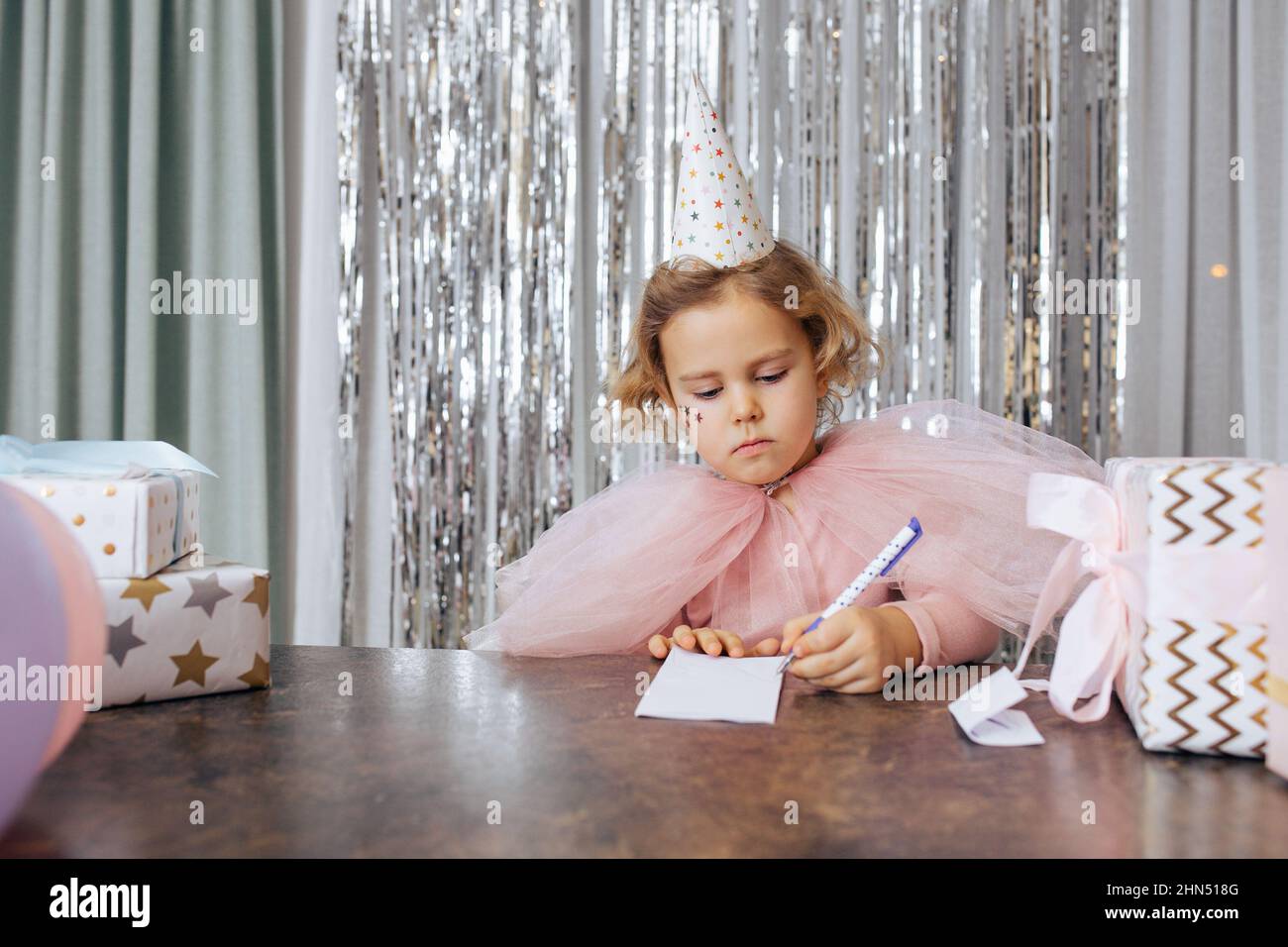 Cute little girl with short curly hair and stars on face in pink poofy dress and birthday cap writing on sheet of paper. Stock Photo