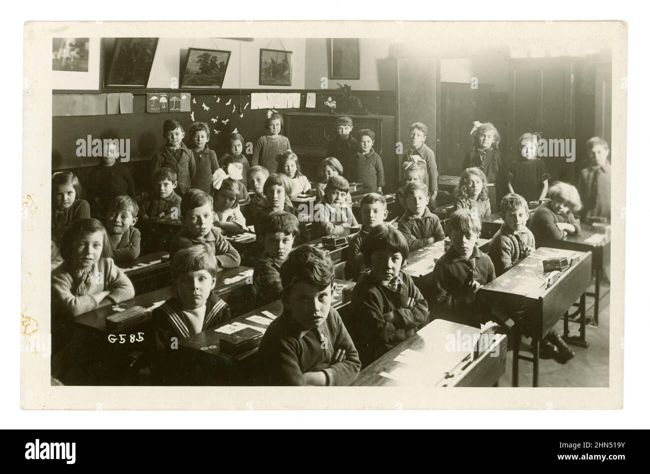 Original early 1920's postcard of serious looking well behaved junior boys and girls sitting at rows of desks, arms folded,  in a junior school classroom, Worthing, Sussex, England, U.K. Stock Photo