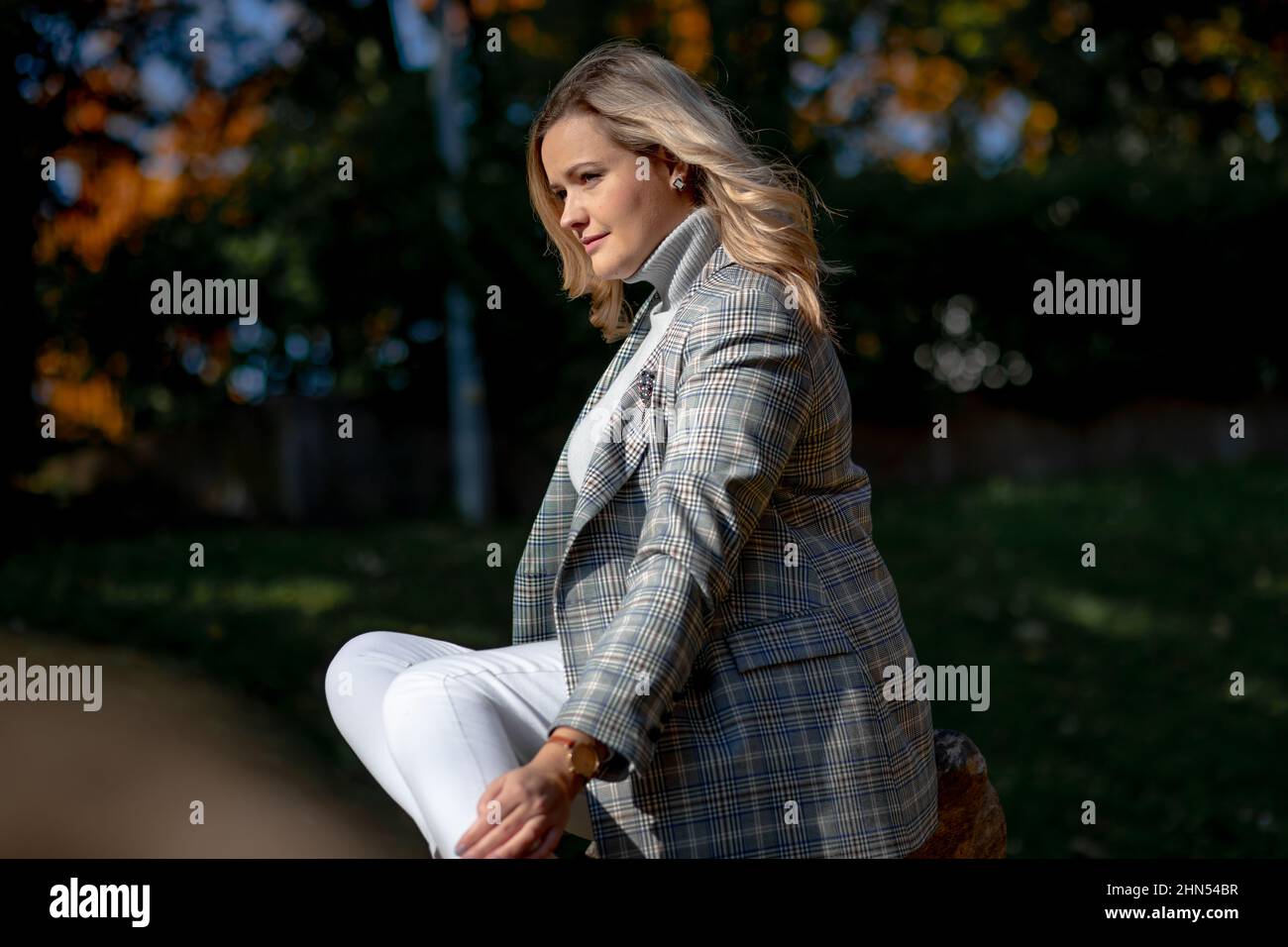 Fashionable smiling middle-aged woman with long wavy fair hair in stylish checkered jacket and roll-neck sweater sitting Stock Photo
