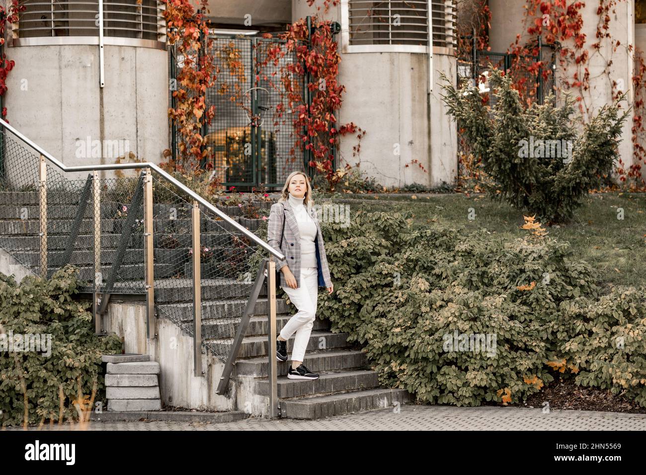 Attractive middle-aged woman in checkered jacket and grey roll-neck sweater going down concrete stairs on autumn day. Stock Photo