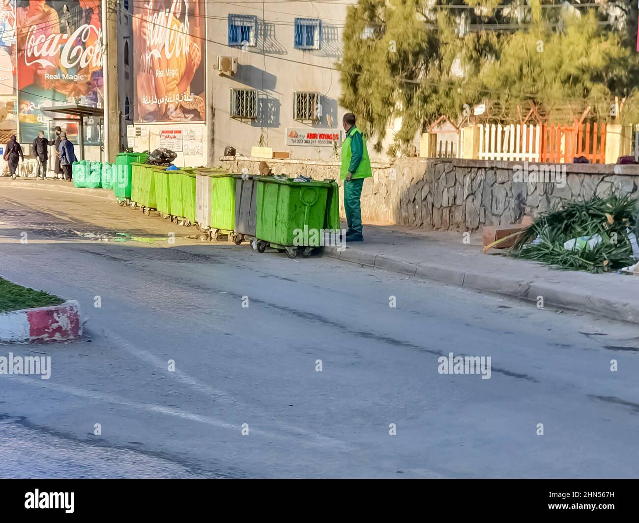 Cheraga, Algiers. Bus station, dumpsters, green bags rubbish palmtree branches, cartons waste. An unrecognizable dustman alone in the sidewalk. Stock Photo