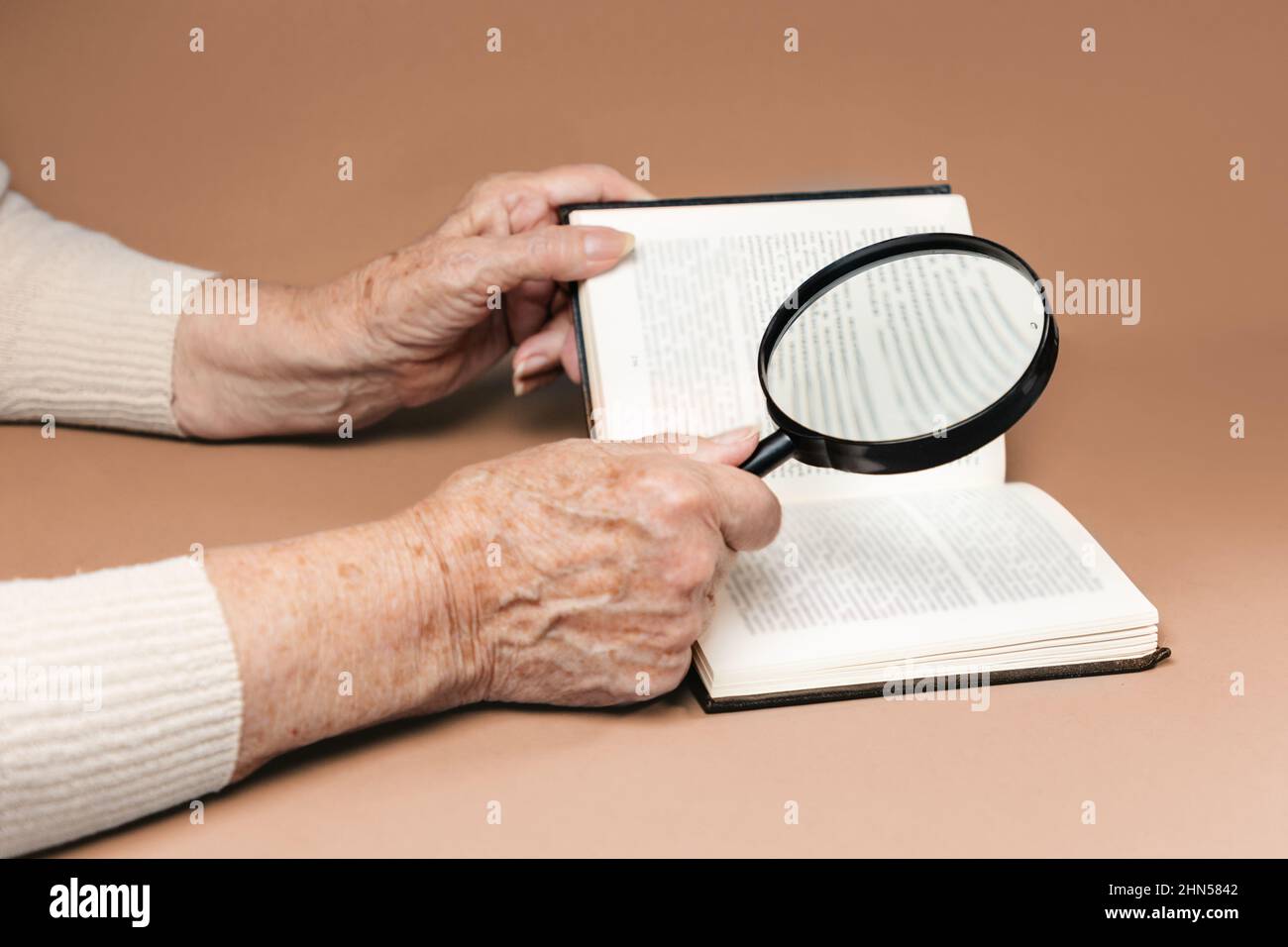 Wrinkled hands of a senior woman are reading a book with magnifier