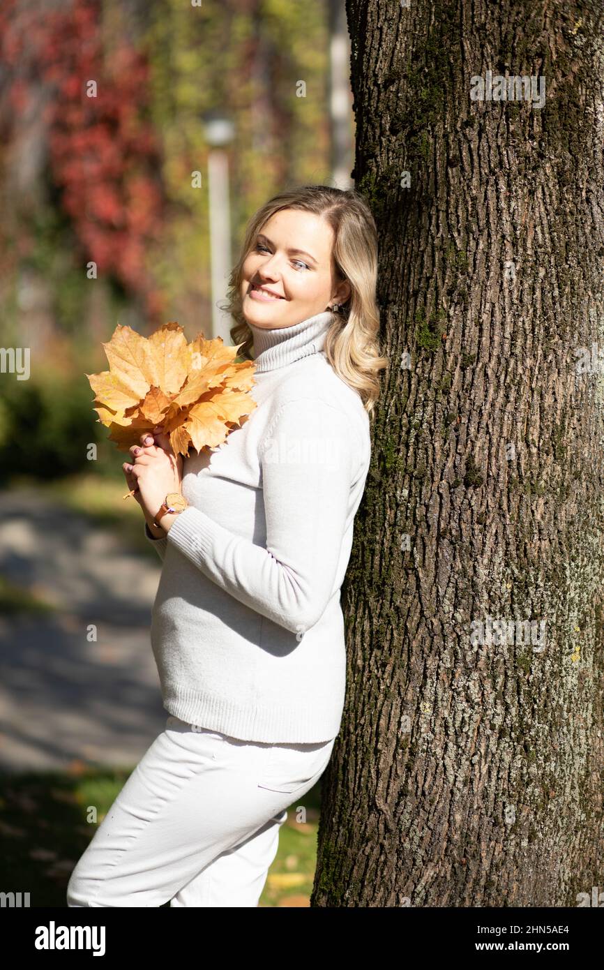 Smiling middle-aged woman with long wavy fair hair in roll-neck sweater holding bouquet of fallen leaves leaning on tree Stock Photo