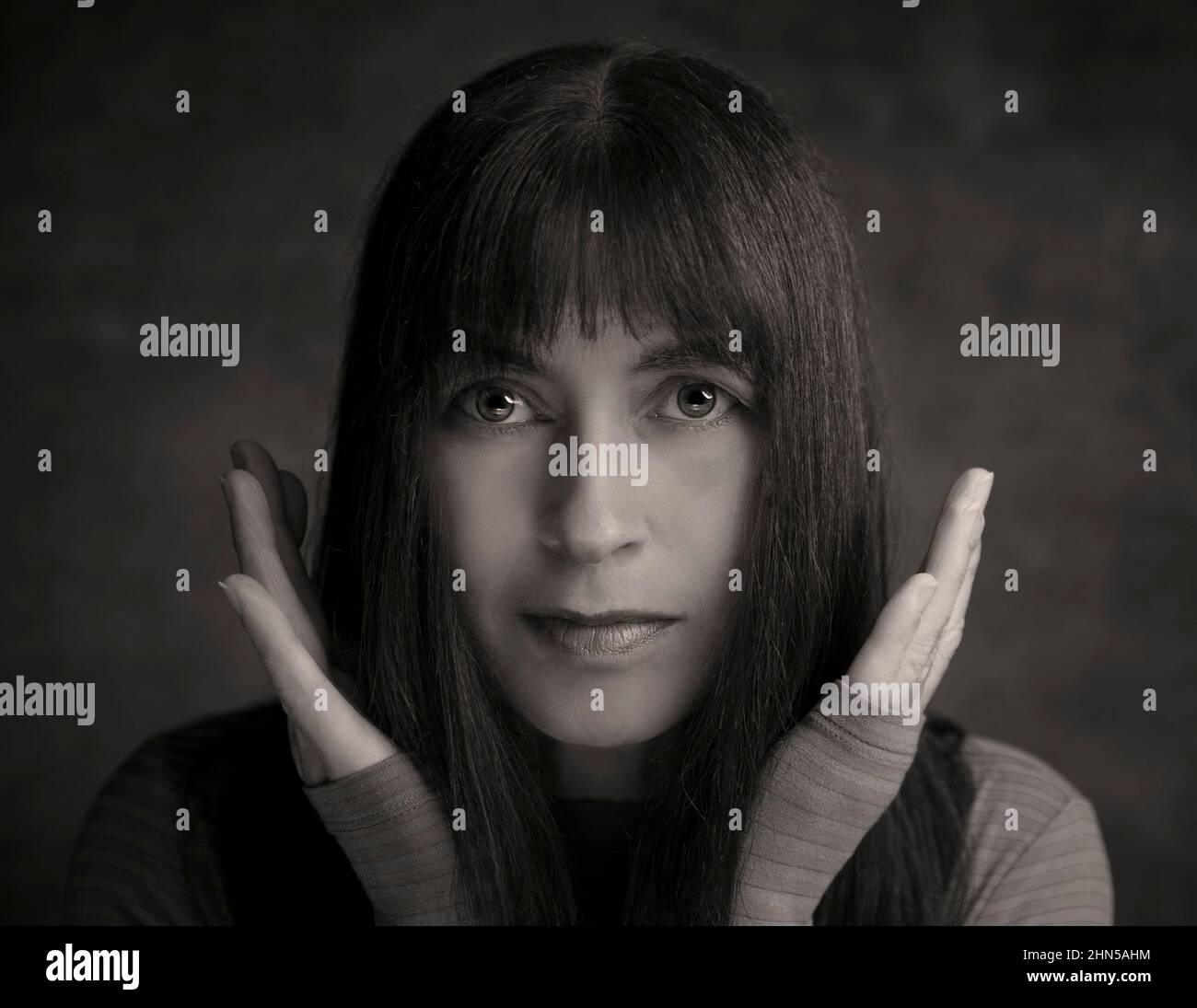 Mono female portraiture: attractive woman looking directly at camera, hands around face. Moody, mysterious portrait of pretty lady with long dark hair. Stock Photo