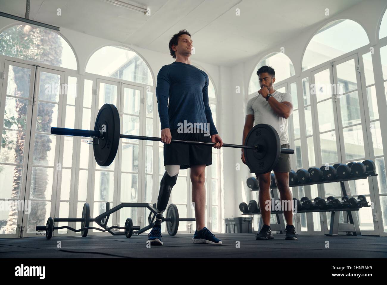 Paralympic athlete weightlifting with his coach in the gym. Man with prosthetic leg being coached by his instructor  Stock Photo
