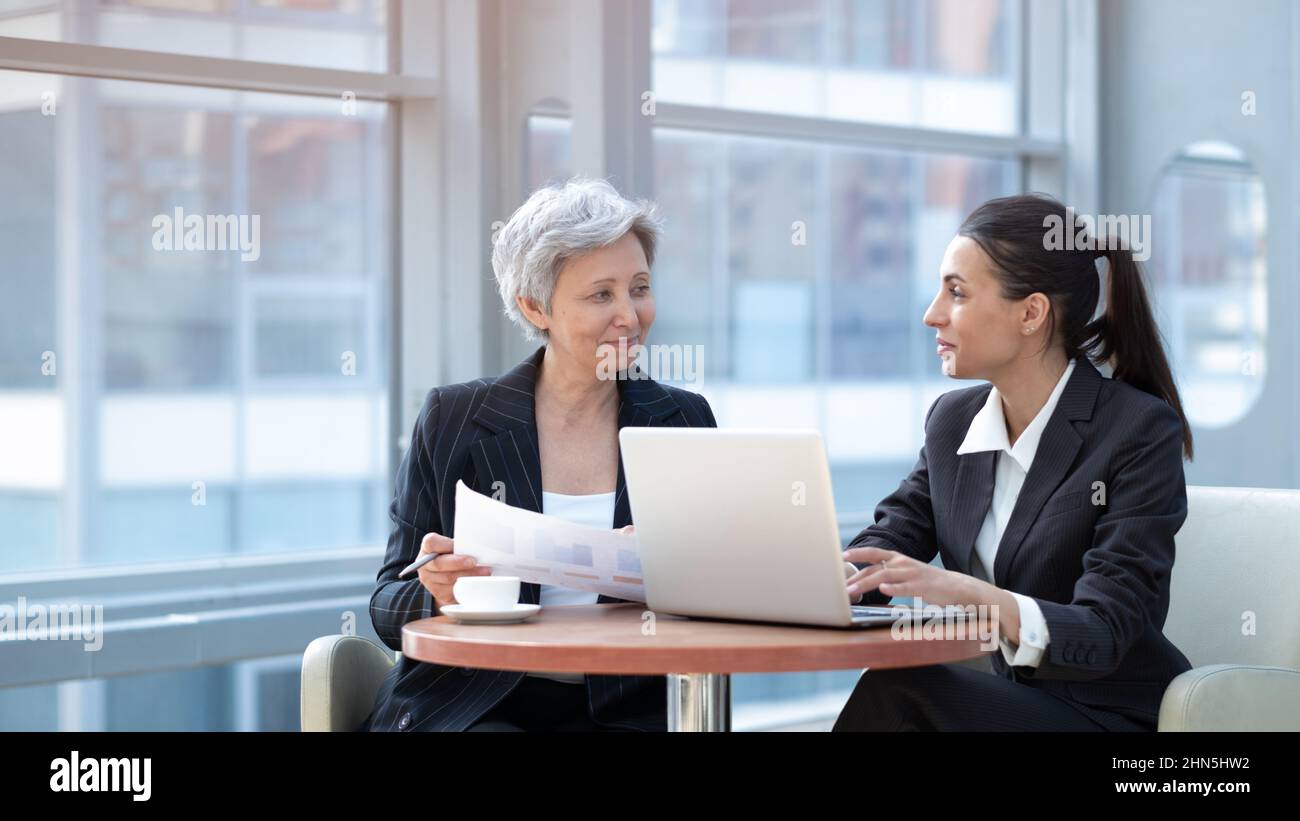 Two women analyzing documents while sitting on a table in office. Woman executives at work in office discussing some paperwork using laptop Stock Photo