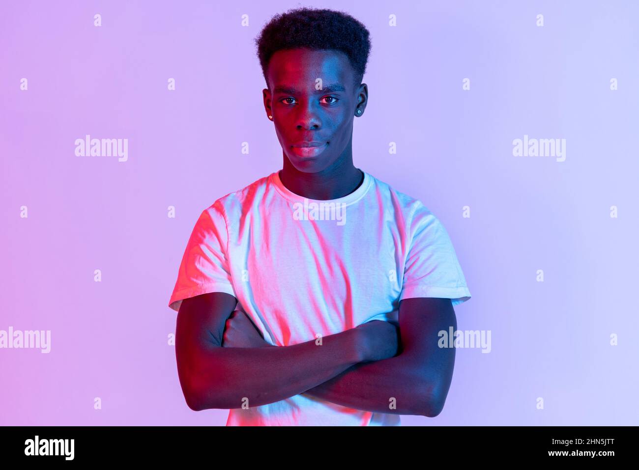Front portrait of a black young man with folded arms, standing in white t-shirt against purple and red lit background Stock Photo