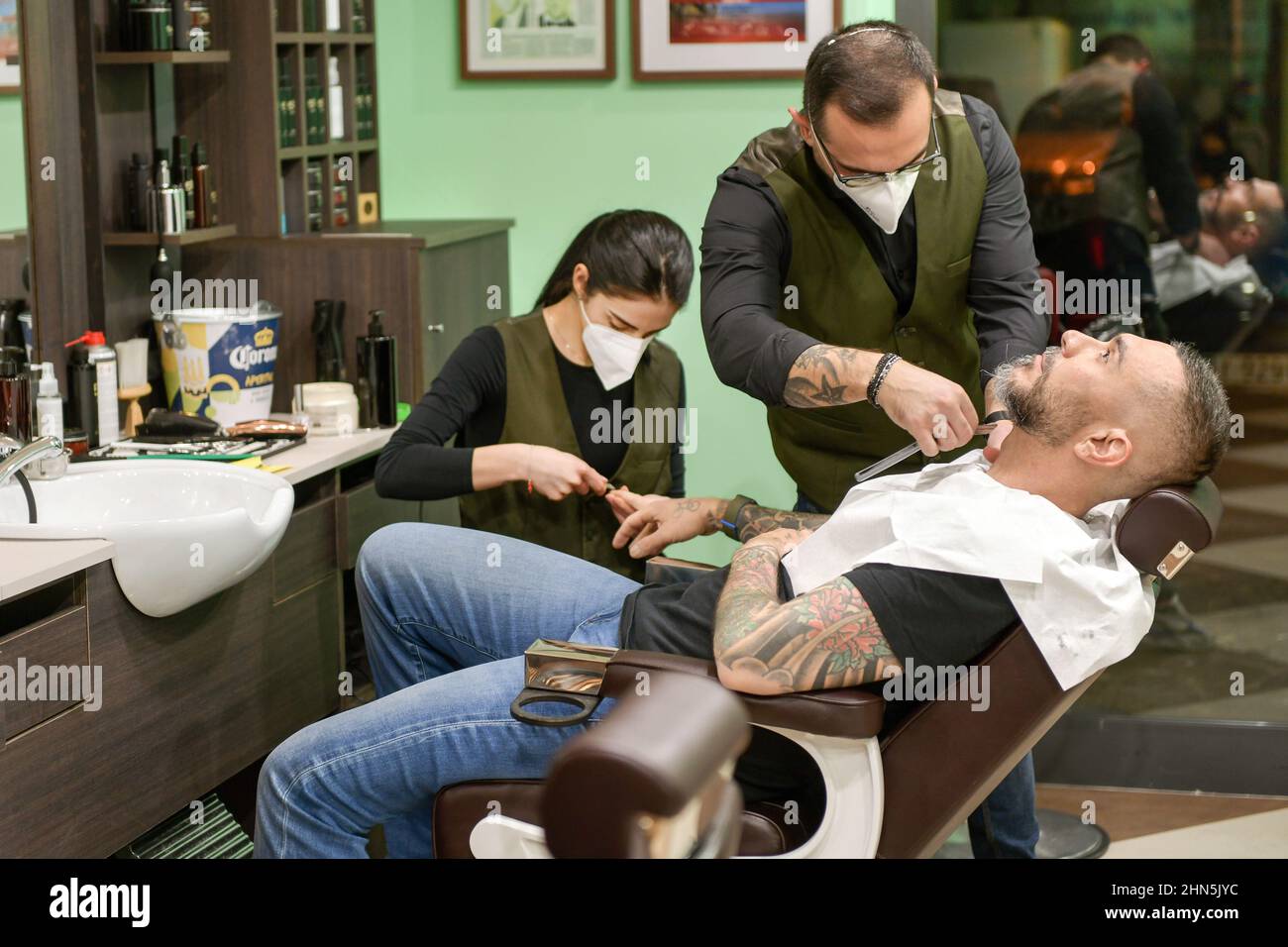 Man with tattoos have a beauty treatment and hair cut in a barbershop or salon with the barber trimming his bear and moustache as the beautician gives Stock Photo