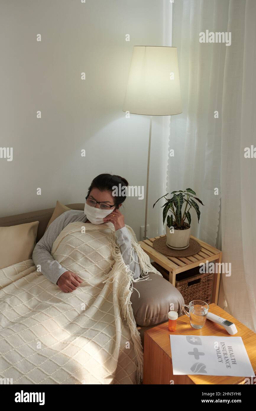 Sick senior woman in medical mask sitting on sofa under warm blanket