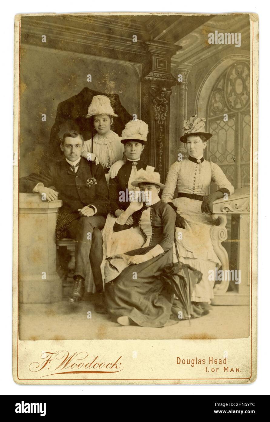 Original, clear, Victorian cabinet card studio portrait of a handsome gentleman wearing a dapper suit, with 4 attractive women, the group are probably tourists. The women are all wearing beautiful dresses and elaborate hats decorated with ostrich feathers in them (the fashion for hat feathers endangered some bird species and led to the setting up of the Royal Society for the Protection of Birds (RSPB) in Britain in 1889), Wonderful painted classic-style backdrop. From the studio of celebrated photographer F. Woodcock, at Douglas Head, Isle of Man, Europe. Circa 1884 Stock Photo