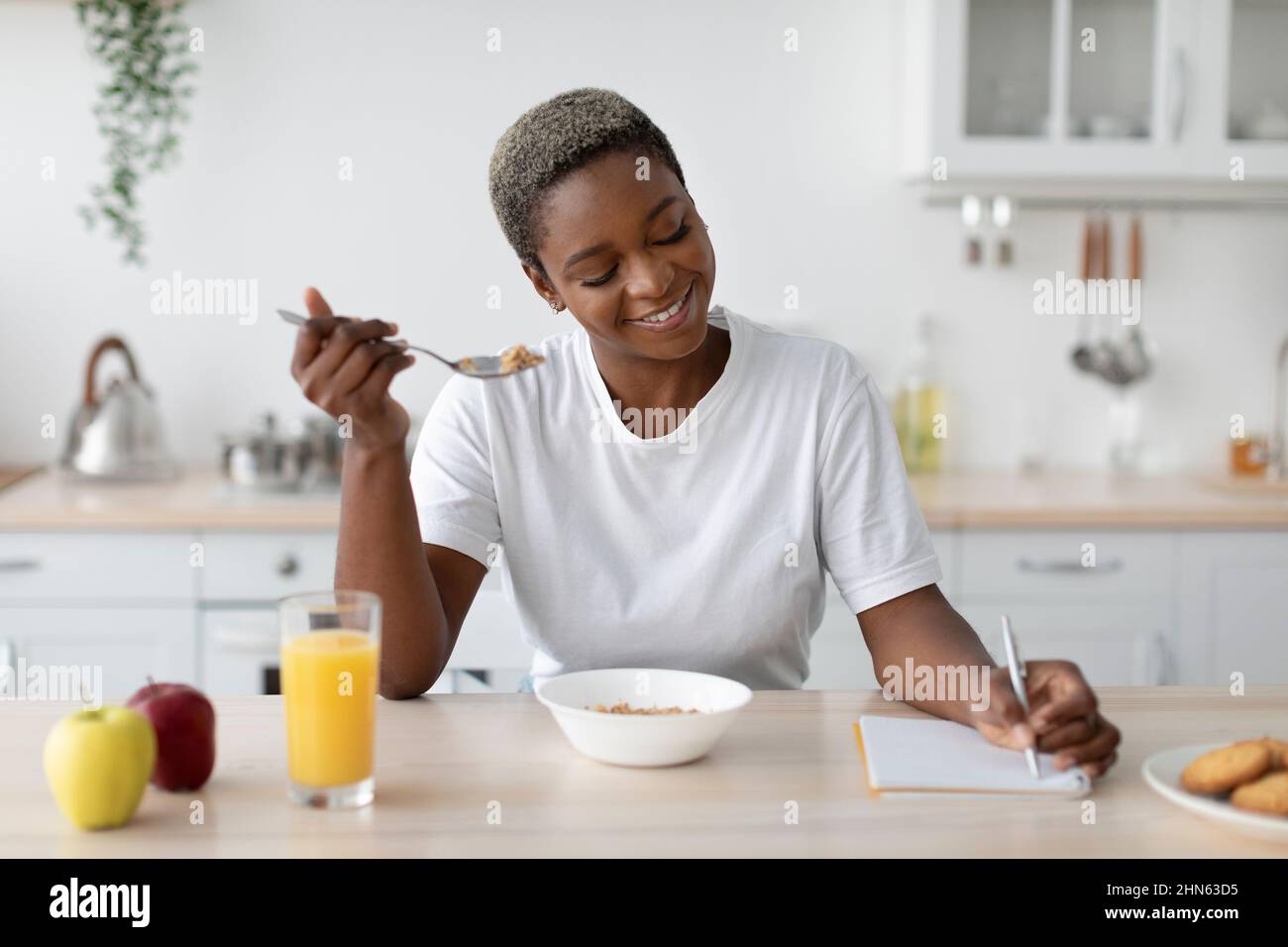 Happy millennial african american female in casual eating porridge, making notes at table Stock Photo