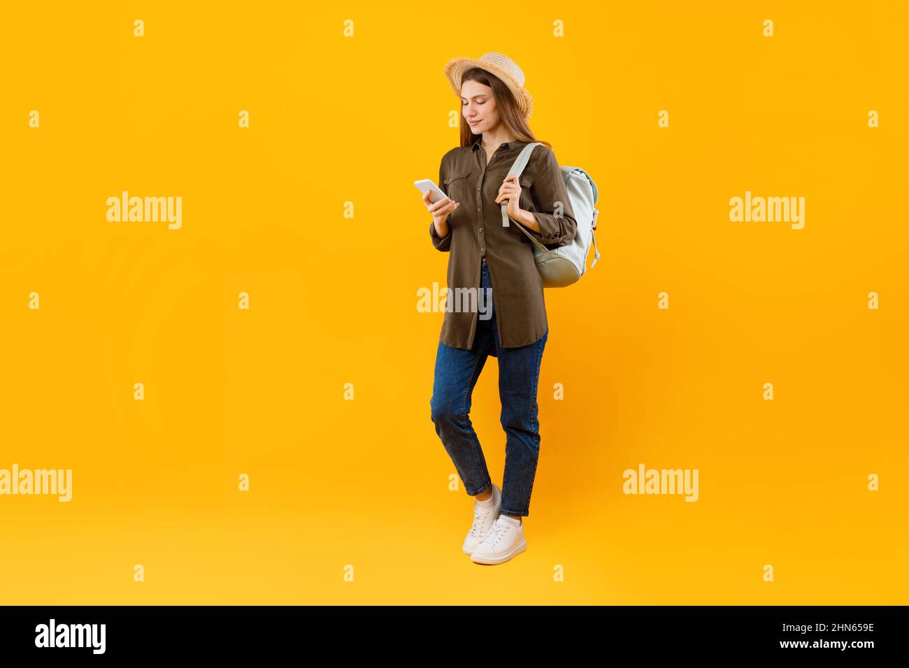 Tourist Lady Using Smartphone Browsing Internet Holding Backpack, Yellow Background Stock Photo
