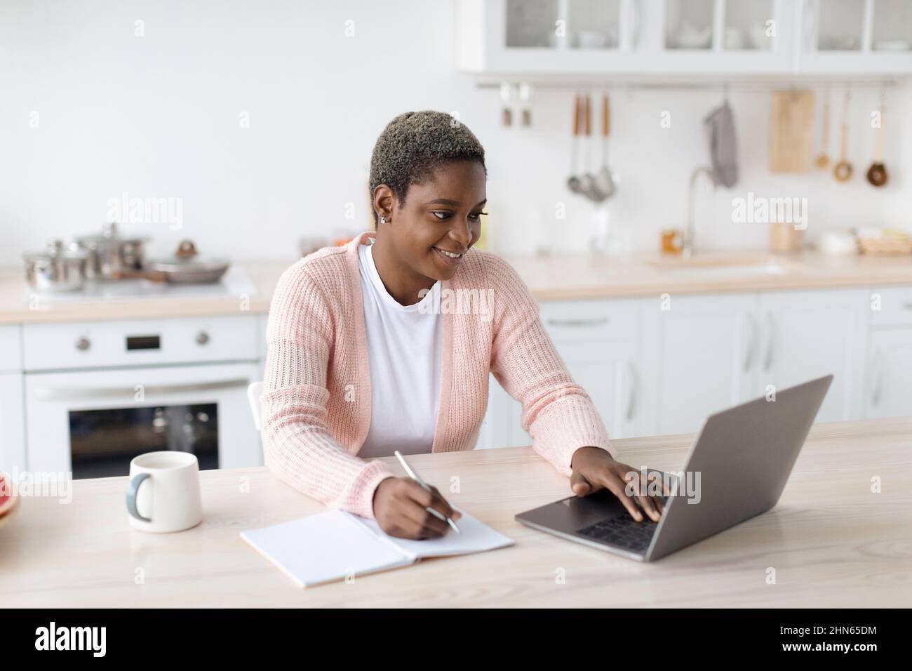Smiling young african american woman in casual working on laptop in minimalist kitchen interior Stock Photo