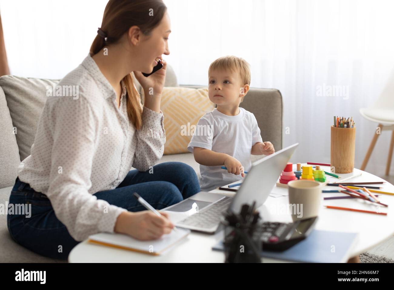 Young busy mother working from home with kid, talking on cellphone and making notes in notepad Stock Photo