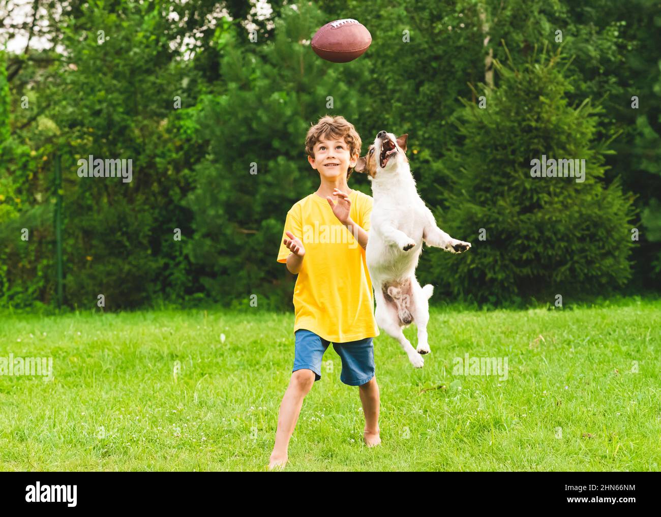 Happy child training to play American football on backyard lawn. Kid and dog catching ball. Stock Photo