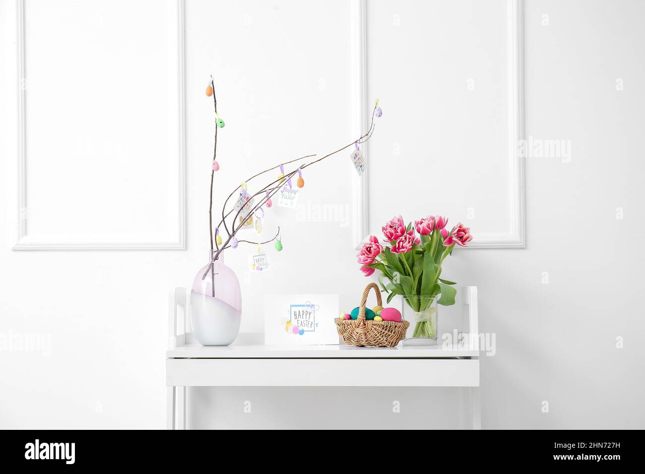 Beautiful Easter composition with basket, painted eggs, flowers and greeting card on table near white wall Stock Photo