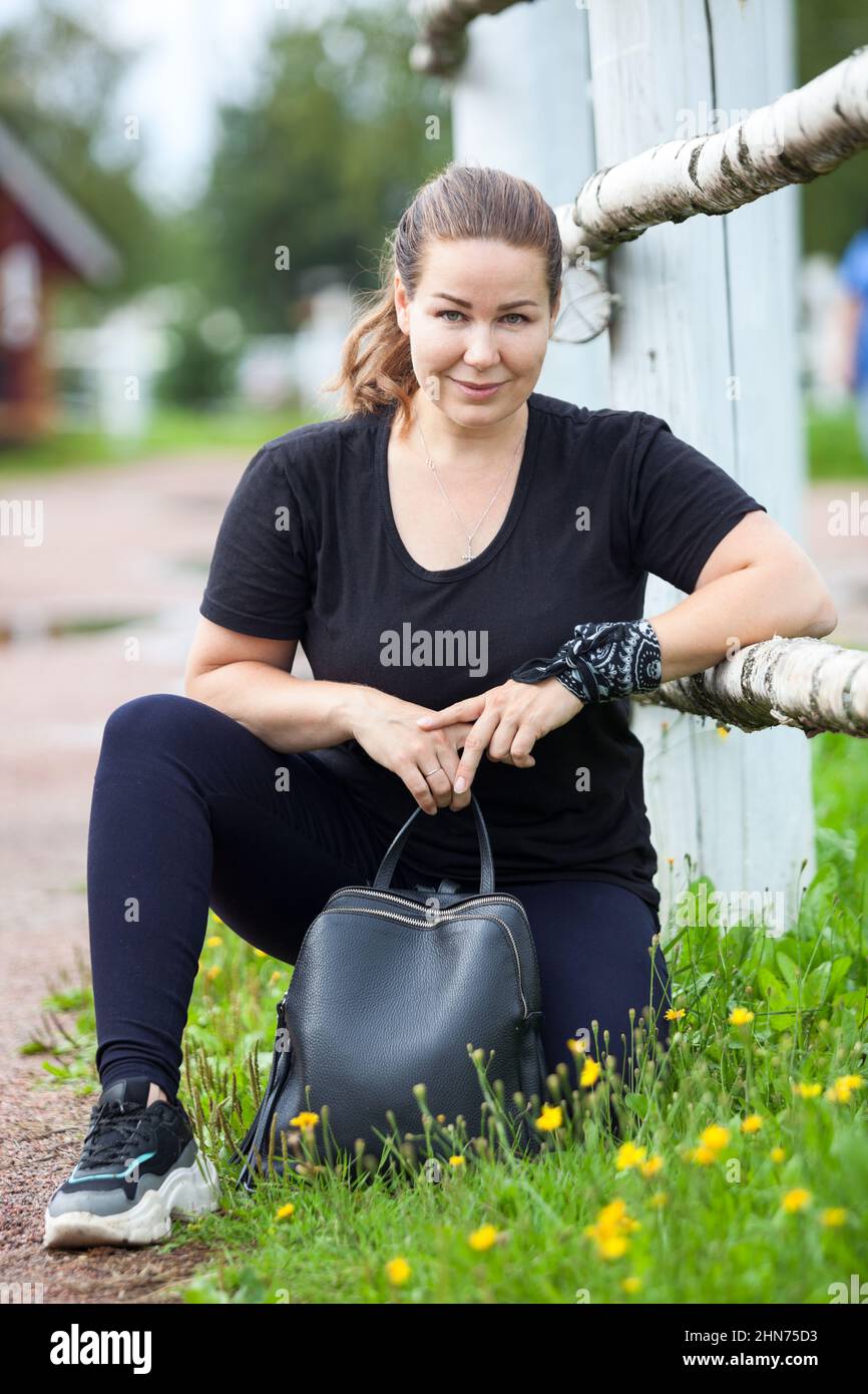 Carefree Caucasian woman walking in summer park, sitting close to fence with black leather dag Stock Photo
