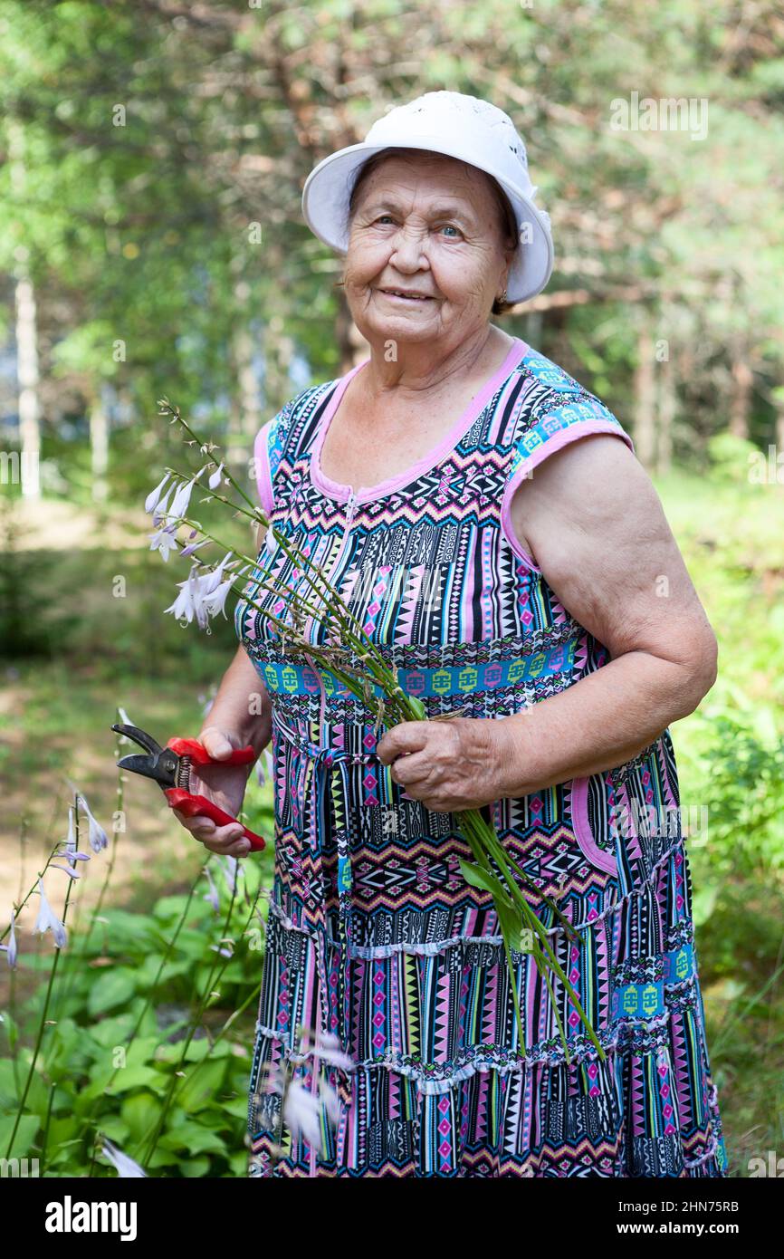 Cheerful old lady with scissors in hands standing in her garden at summer season, Caucasian woman Stock Photo