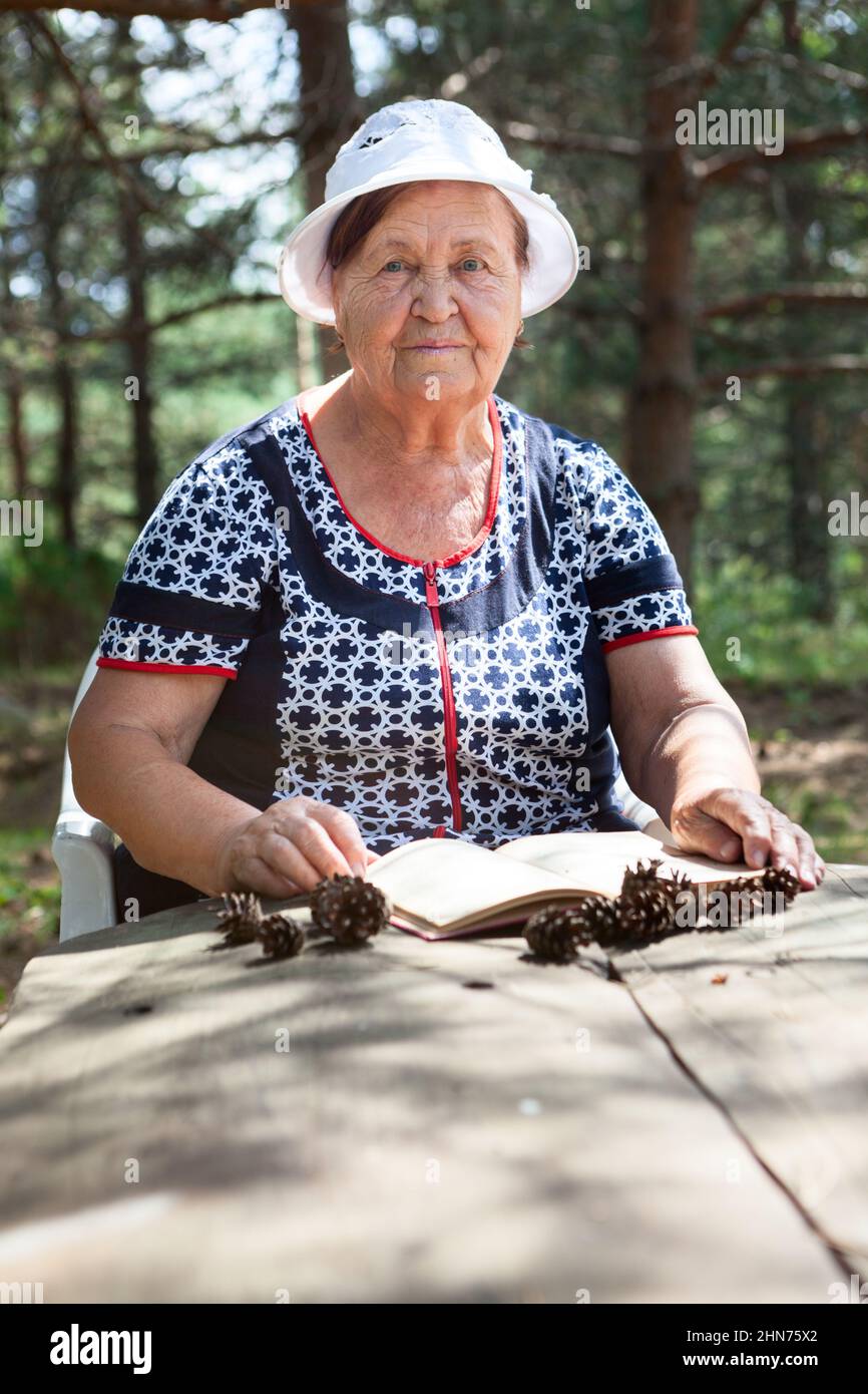 Old lady reading book sitting at the wooden table in evergreen forest, looking at camera Stock Photo