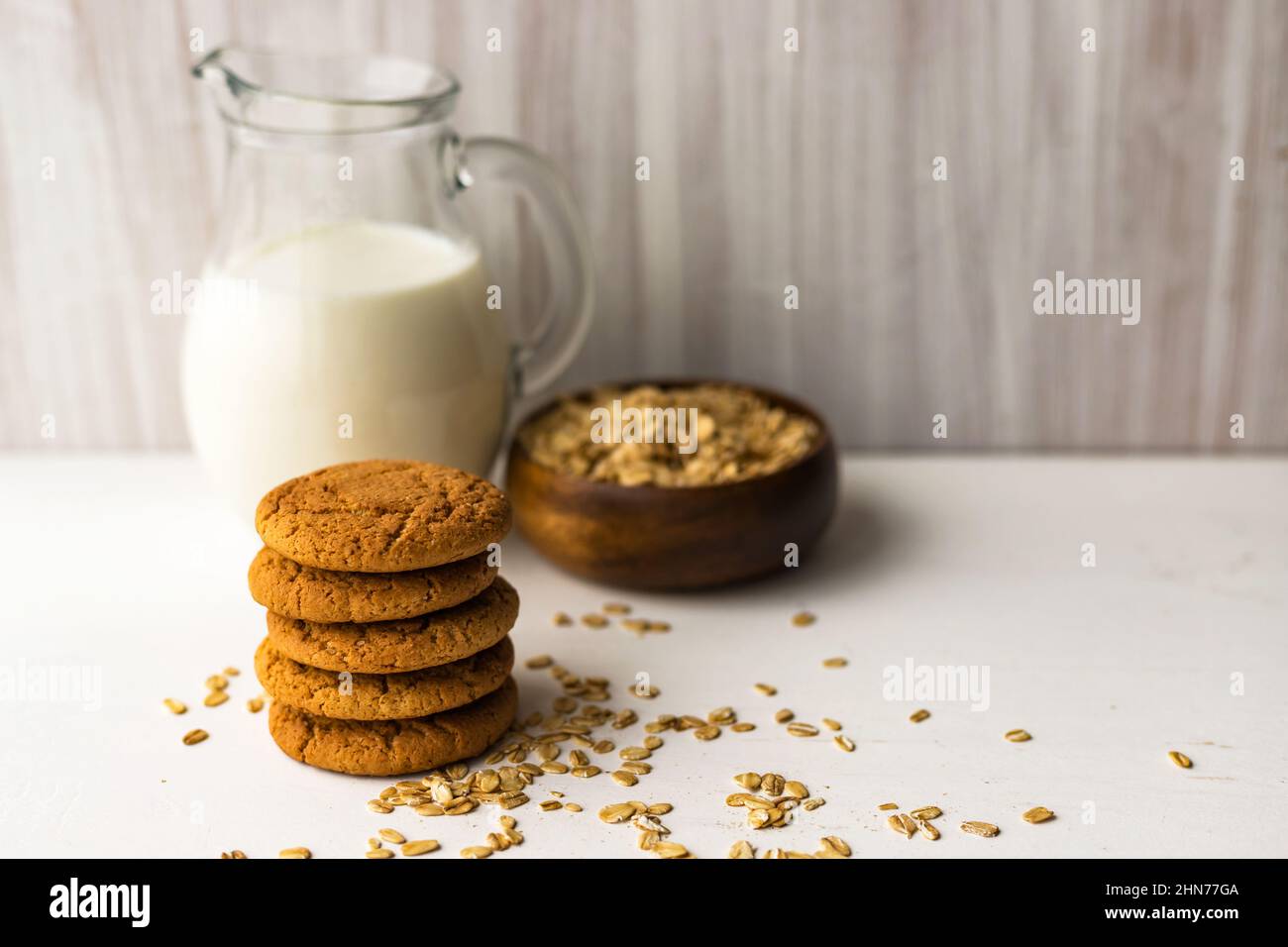 Heap oatmeal cookies and oat cereal flakes bowl on white table on milk glass jug background Stock Photo
