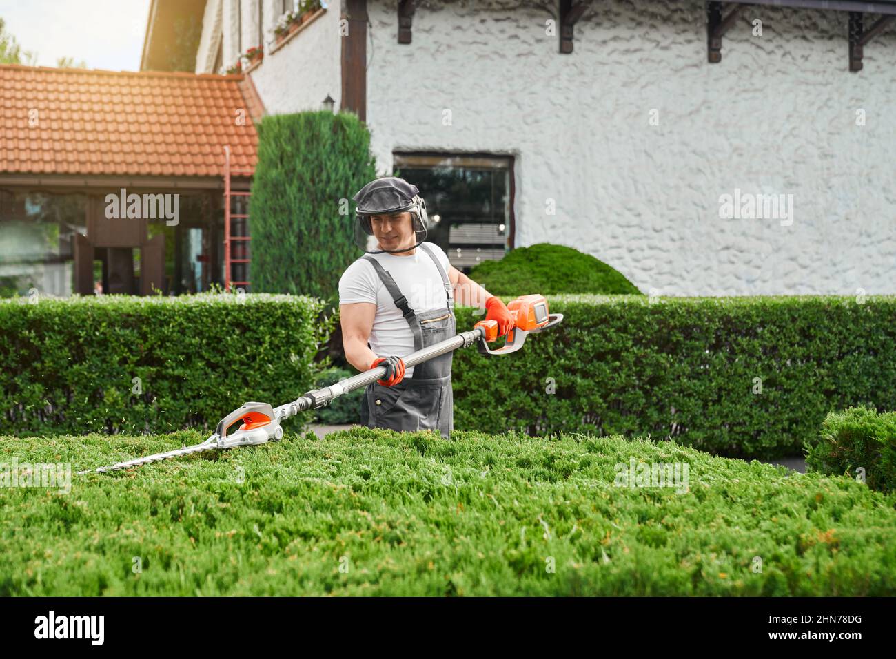 Caucasian worker in uniform, gloves and safety mask cutting overgrown bushes with petrol hedge