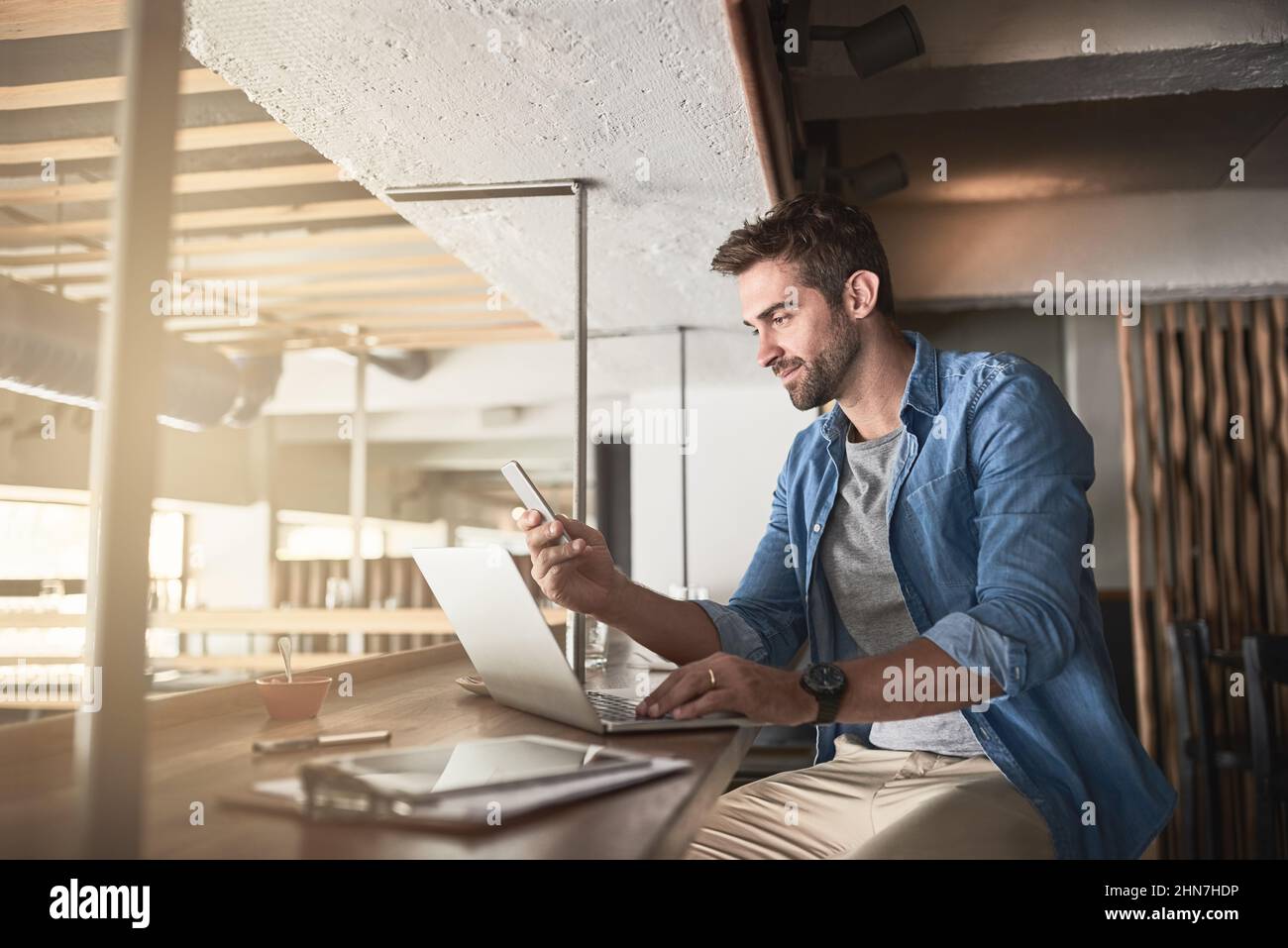 The hard work that goes into running a cafe. Shot of a handsome young man using a laptop and phone in a coffee shop. Stock Photo